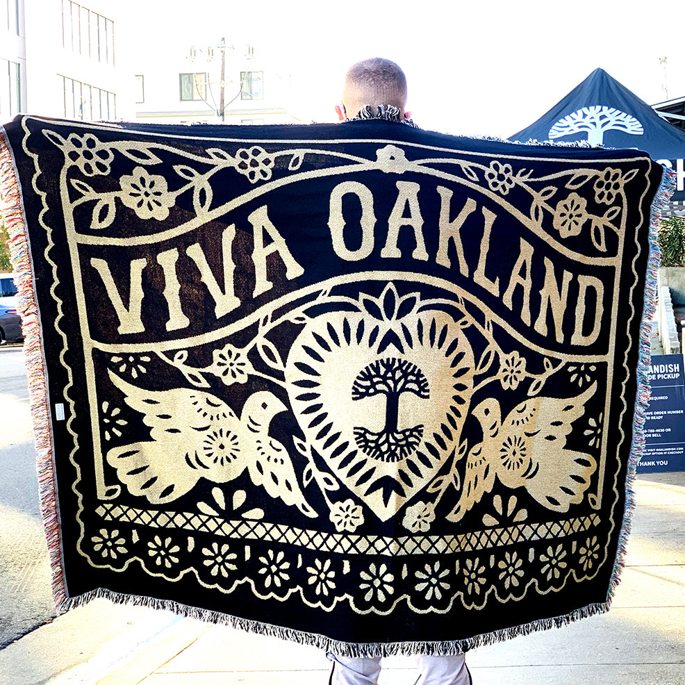 A person holds up the Viva Oakland Throw by Oaklandish, a large black and cream woven cotton blanket that reads "Viva Oakland" in bold text. It features two birds and a heart encircling a tree, with floral patterns adorning the top and bottom. The person is standing outdoors with buildings and a tent visible in the background.