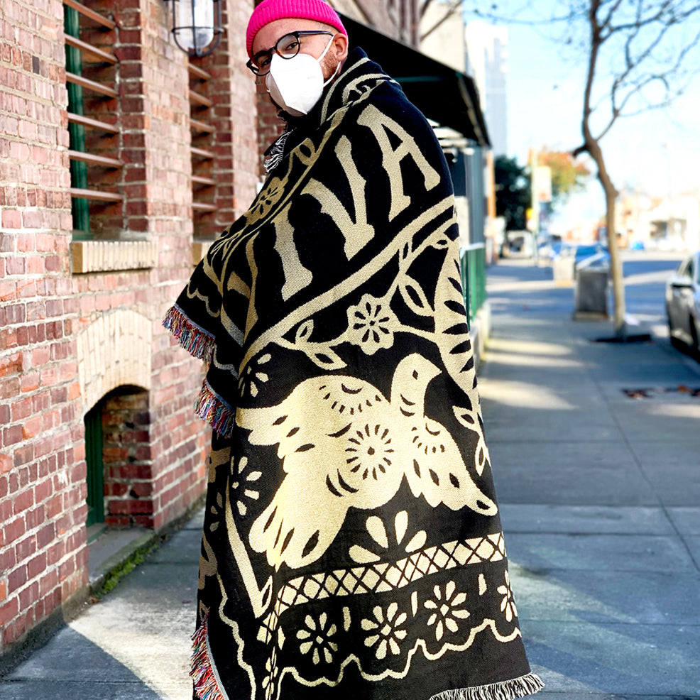 A person in a pink beanie and glasses, wearing a white face mask, stands on a sidewalk next to a brick building. They are wrapped in the Viva Oakland Throw by Oaklandish, which is a large black and beige woven blanket featuring a bird and floral design made from recycled fibers. The background shows a street scene with trees and buildings.