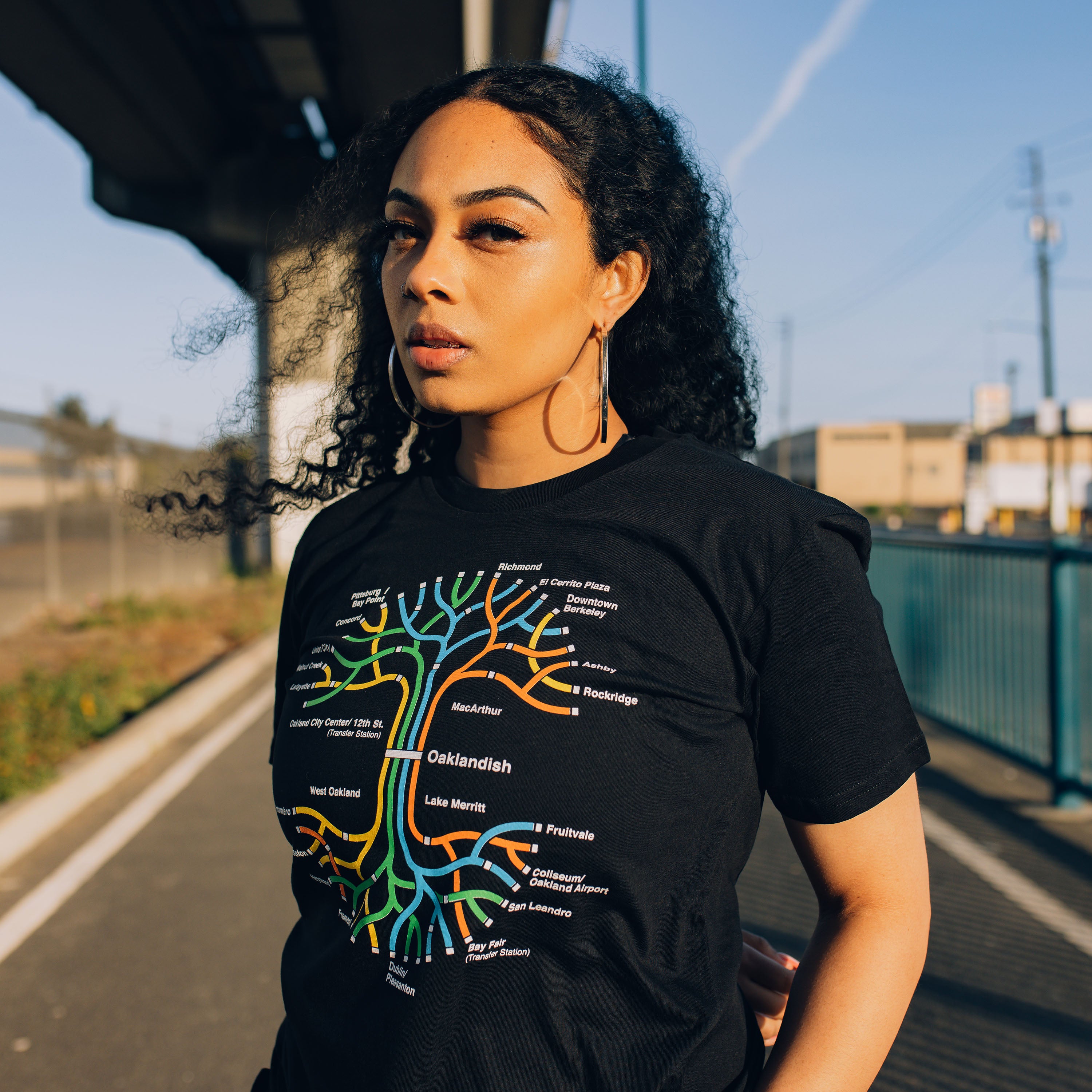 A woman stands outdoors on a sunny day, wearing the BART Tree Tee by Oaklandish. The black T-shirt, showcasing a colorful tree-like graphic with text, is a 100% cotton classic fit. She has long hair and is in an urban setting surrounded by buildings, a rail track, and a blue fence. The sky is mostly clear with a few wisps of clouds.