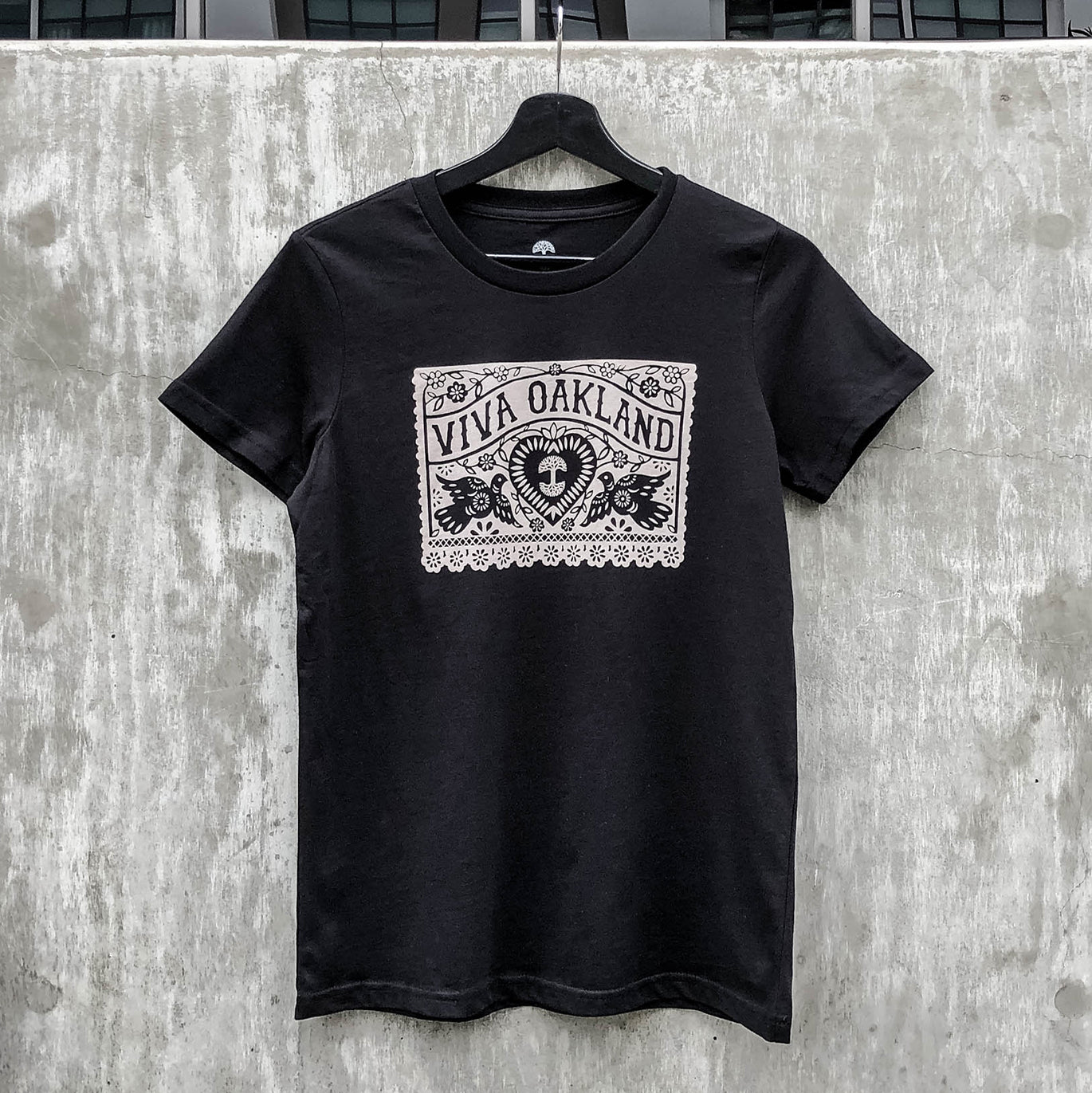 A black Women's Viva Oakland Tee from Oaklandish, made of 100% cotton, with a white design hangs on a black hanger against a concrete wall. The classic fit T-shirt features ornate floral patterns surrounding a central emblem with the text "VIVA OAKLAND" at the top and a stylized image of a heart with rays beneath it.