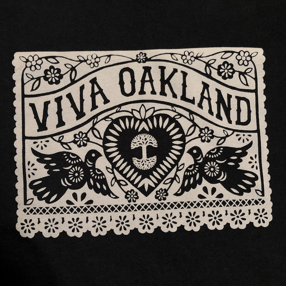 The Women's Viva Oakland Tee by Oaklandish showcases a striking black and white design that mimics cut-paper artwork. Prominently displaying "VIVA OAKLAND" in bold letters, it features floral elements, two birds facing each other, and a heart with a tree at its center. A lacy-edged border adds to the decorative appeal of this 100% cotton classic fit t-shirt.