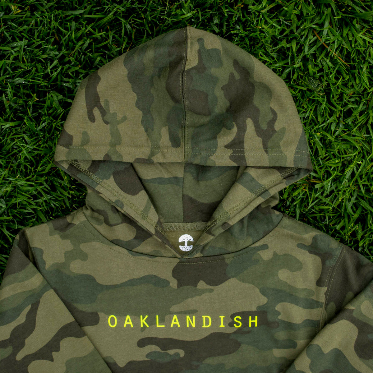 An image of the Youth Classic Hoodie by Oaklandish in a camouflage pattern, featuring "OAKLANDISH" in bright yellow letters. The kids size hoodie is laid flat on a grassy background, with a small tree logo visible inside the hood.