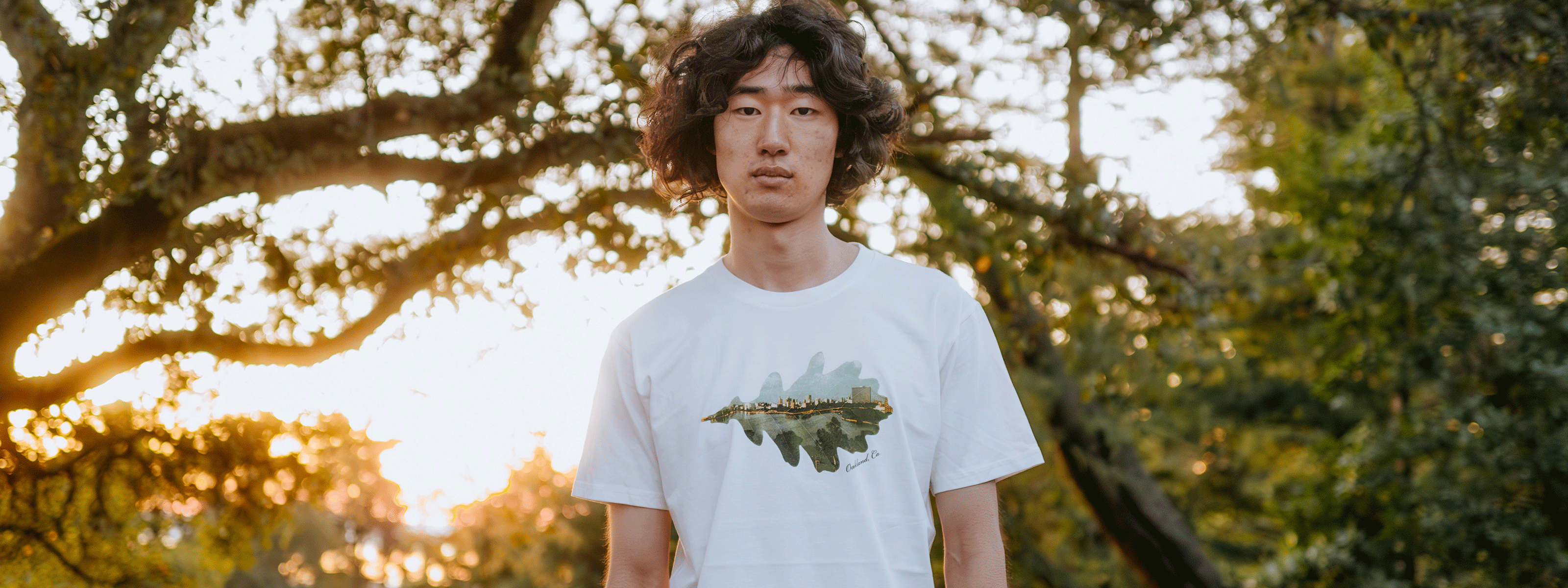 A person with curly hair stands outdoors wearing a white T-shirt with a nature-themed design. The background features trees and a sunset, creating a warm, serene atmosphere.