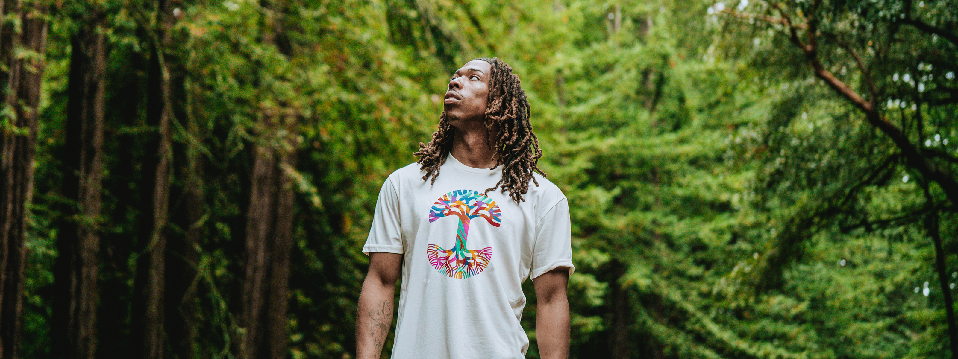 A person wearing a black T-shirt with “OAKLANDISH” in bold white letters and a colorful abstract design on the back, standing outdoors with trees and misty hills in the background.