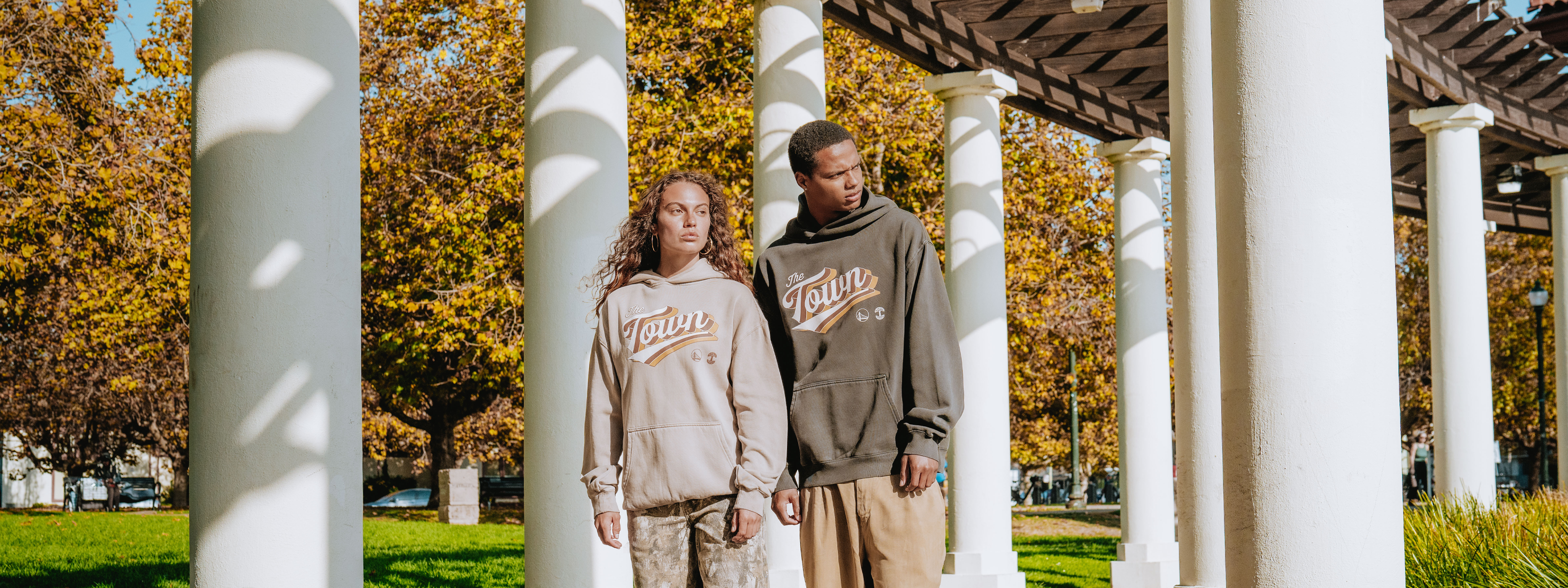 Two people sit close together wearing matching printed sweatshirts and beige pants, with modern glass buildings and a clear blue sky in the background. Their expressions are calm and confident.