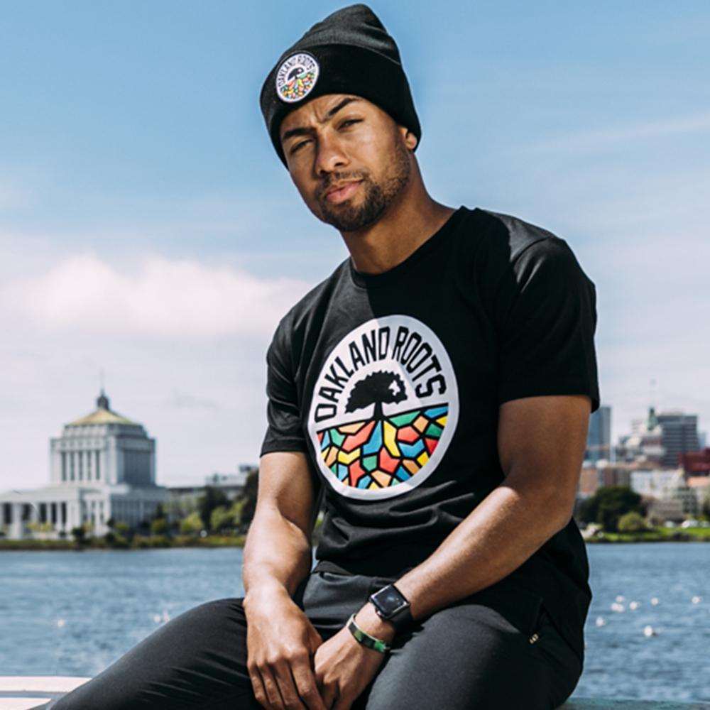 A man dressed in a black "Oakland Roots" T-shirt and an "Oakland Roots SC Cuff Beanie," both displaying the Oakland Roots logo, poses in front of a waterfront with city buildings and a domed structure visible in the background. He has a watch on his left wrist and looks calmly towards the camera.