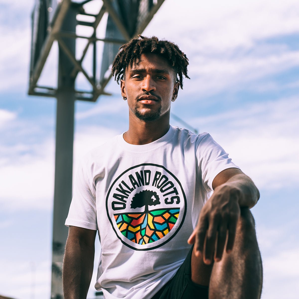 A man with short, curly hair is wearing an Oakland Roots SC Classic Tee featuring a vibrant "Oakland Roots" logo. He is posing outdoors in front of a metal structure with a blue sky and some clouds in the background. The sports club's logo displays an afro silhouette with extended roots in vivid colors.