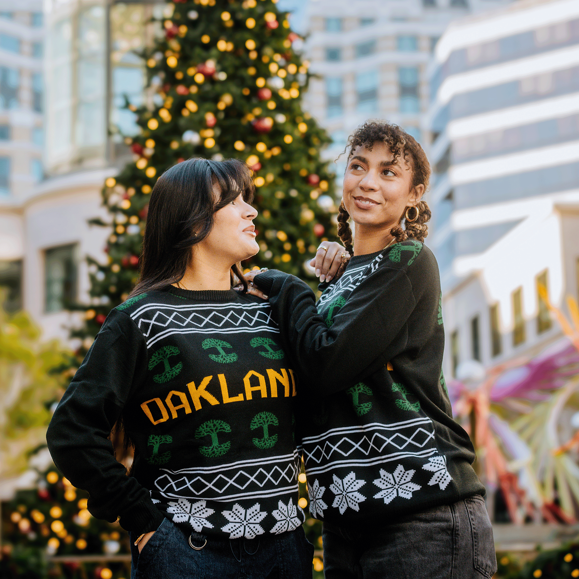 Two people pose in front of a festive Christmas tree outdoors, wearing matching Oaklandish 2024 Ugly Sweaters. These black knit sweaters are decorated with white snowflakes and the word "OAKLAND" in yellow. Behind them towers a modern building with expansive windows. They share warm smiles as one gazes affectionately at the other.