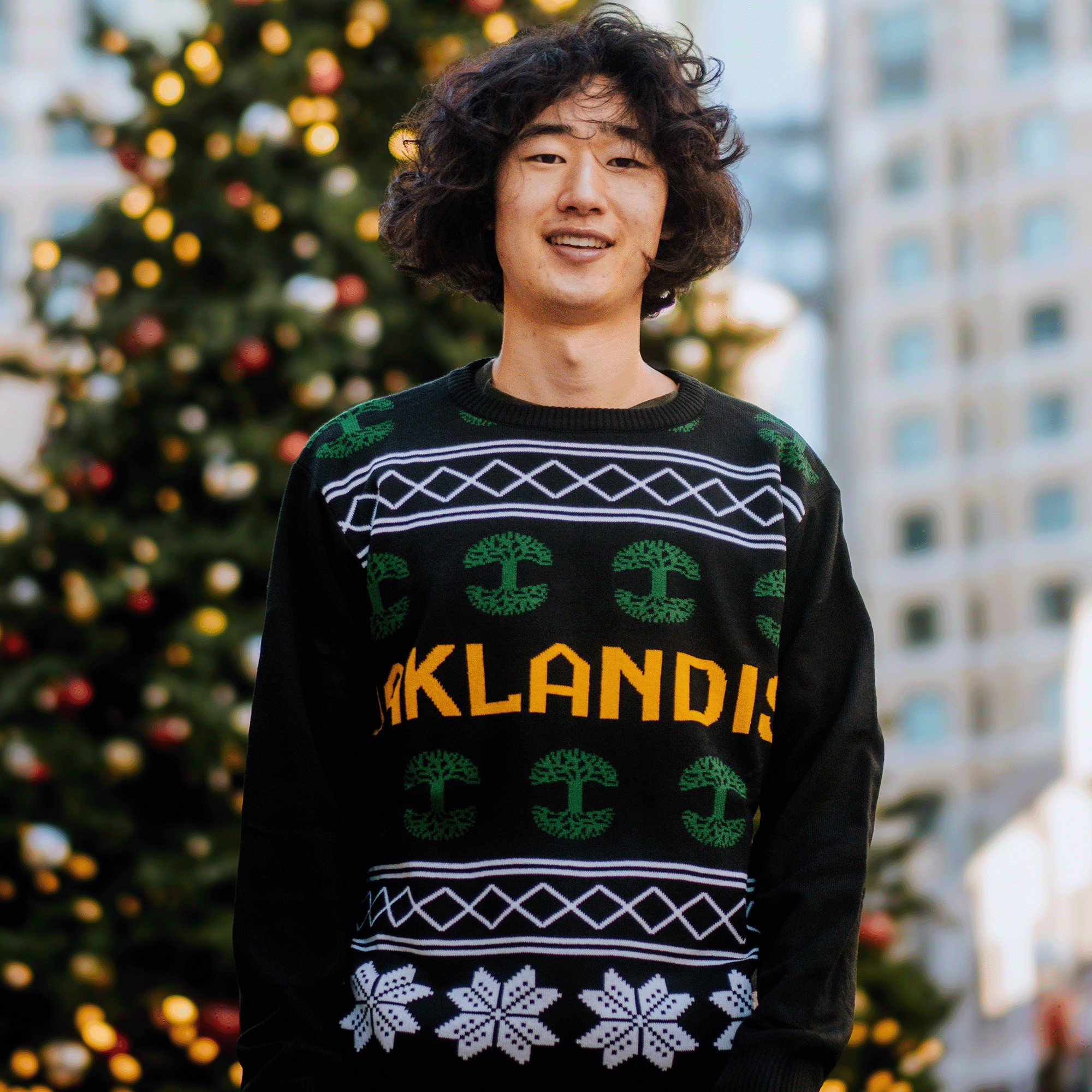 A person with curly hair smiles while standing in front of a decorated Christmas tree, wearing the festive Oaklandish 2024 Ugly Sweater, a black knit sweater adorned with green designs and white snowflakes, featuring the brand name "OAKLANDISH." Buildings can be seen in the blurred background.