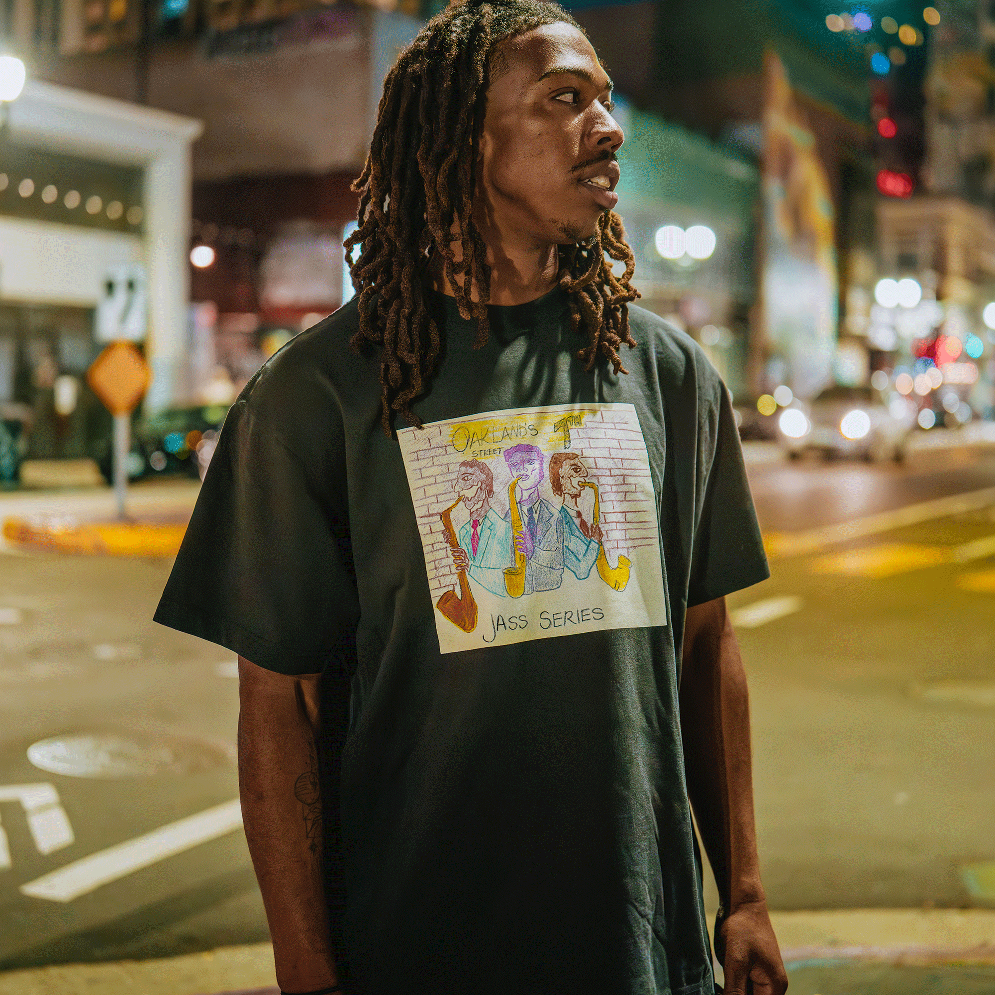 A man with long dreadlocks stands on a city street at night, wearing Oaklandish’s 7th St. Players Tee—a classic fit black t-shirt made from 100% cotton, featuring a colorful “Jass Series” cartoon graphic amid bright city lights.
