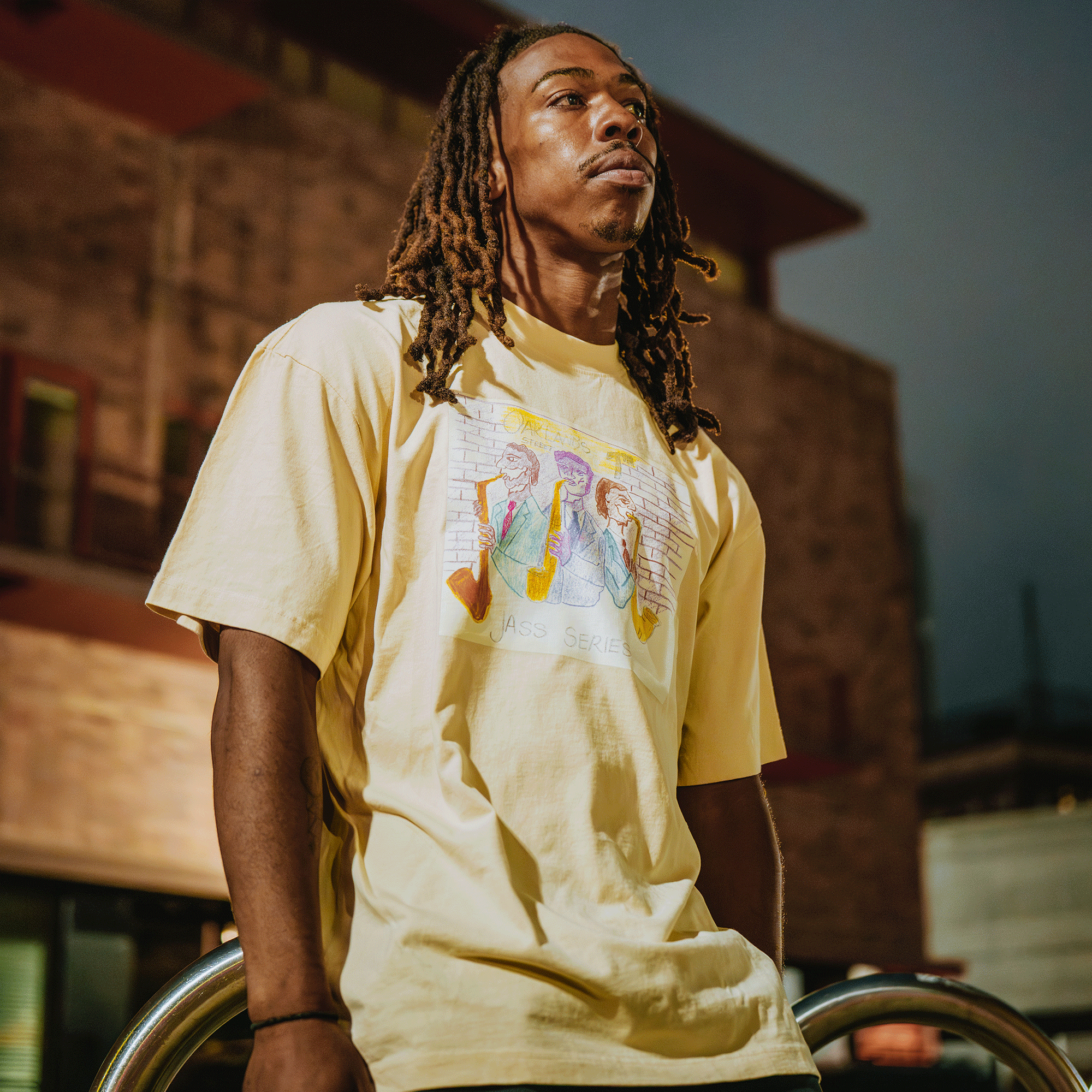 A man with long brown dreadlocks stands at dusk, wearing the Oaklandish 7th St. Players Tee— a natural 100% cotton yellow classic fit shirt featuring an illustrated graphic of musicians—while gazing thoughtfully against urban brick buildings.