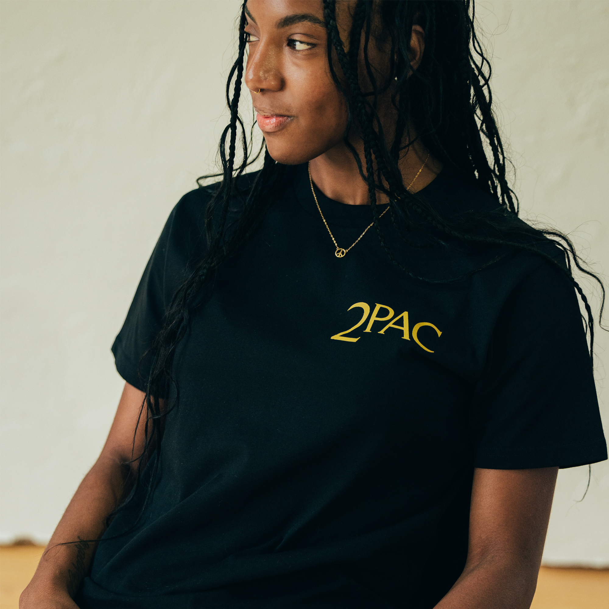 A person with long braided hair is wearing an Against the World Tee, showcasing their deep connection to hip-hop culture. Seated against a neutral background, they look to the side with a slight smile, while an Oaklandish pendant necklace rests around their neck.