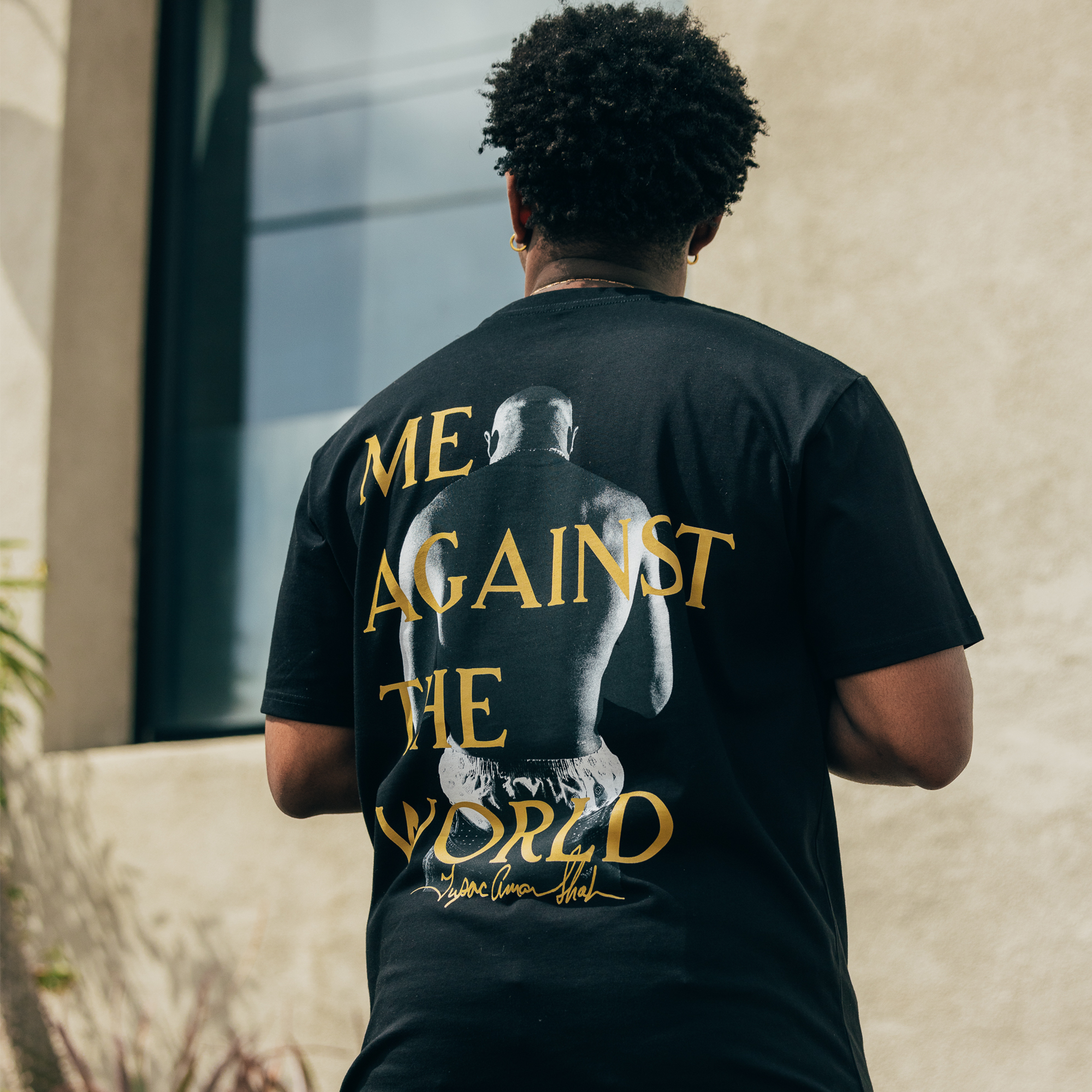 A person with curly hair, seen from behind, is wearing the "Against the World Tee" by Oaklandish. The black T-shirt features yellow text reading "ME AGAINST THE WORLD," accompanied by an image of a boxer. This hip-hop-inspired scene unfolds outdoors near a beige building with a large window, surrounded by plants, capturing an Oaklandish vibe.