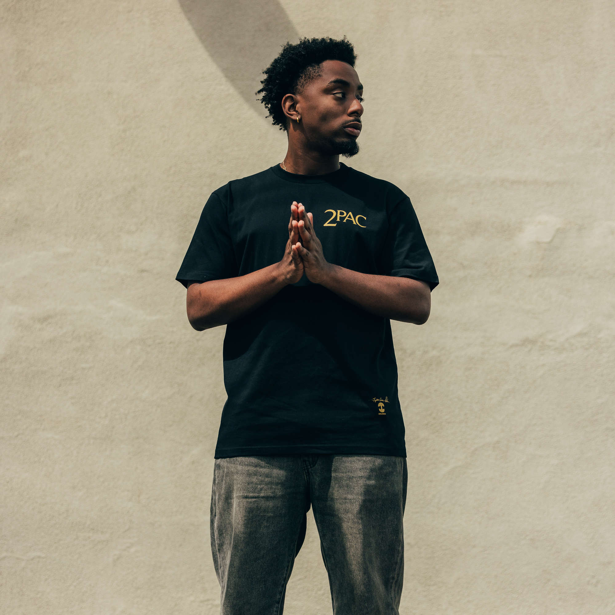 A man stands against a beige wall, donned in a black "Against the World Tee" by Oaklandish and gray jeans, epitomizing hip-hop vibes. He gazes to his right, hands clasped before his chest, as the lighting casts an Oaklandish shadow behind him.