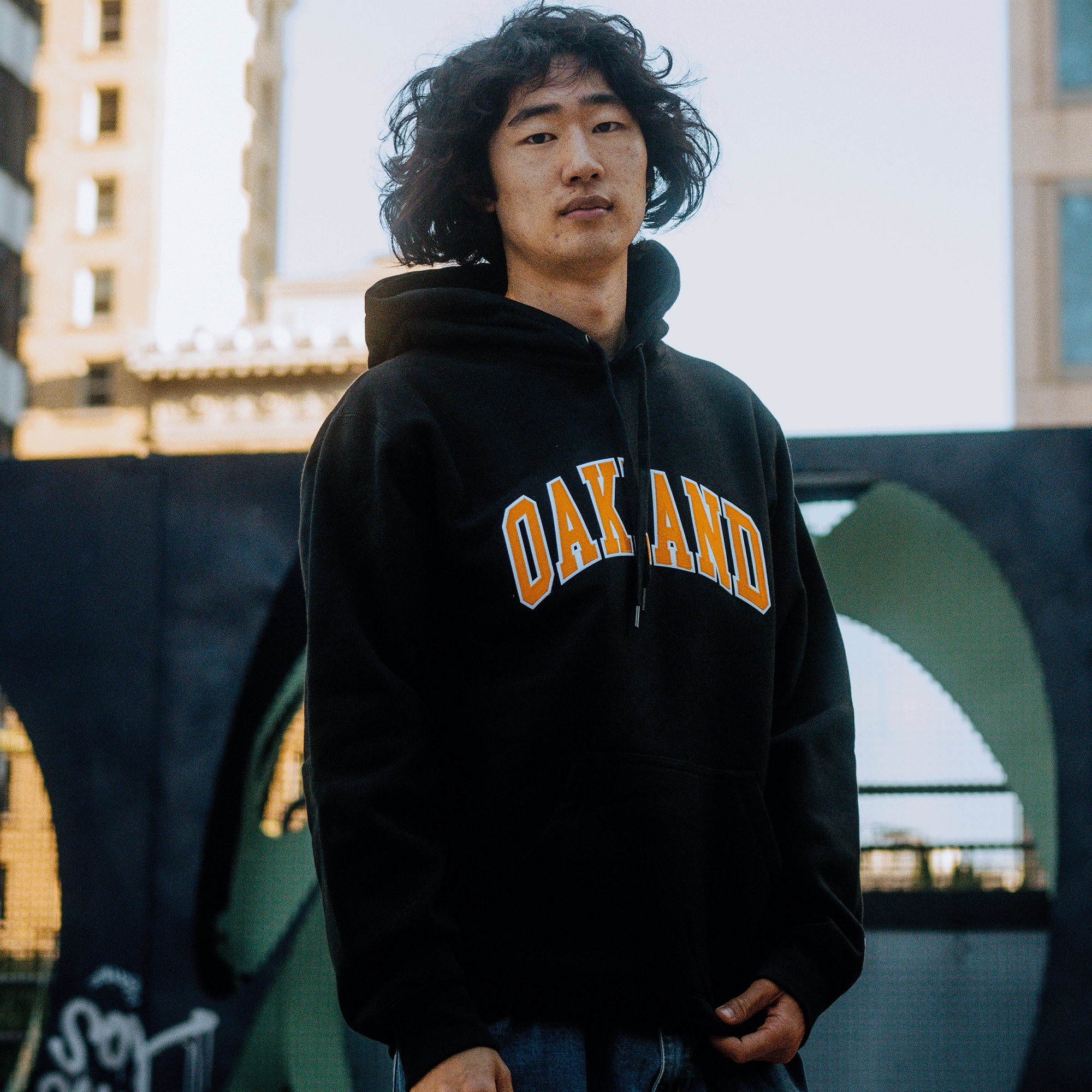 A person with long, wavy hair stands outdoors in an Alma Mater Heavy Hoodie by Oaklandish, featuring the word "OAKLAND" in vibrant yellow text. They appear serious against the backdrop of towering buildings and a curved structure under a clear, sunny sky.