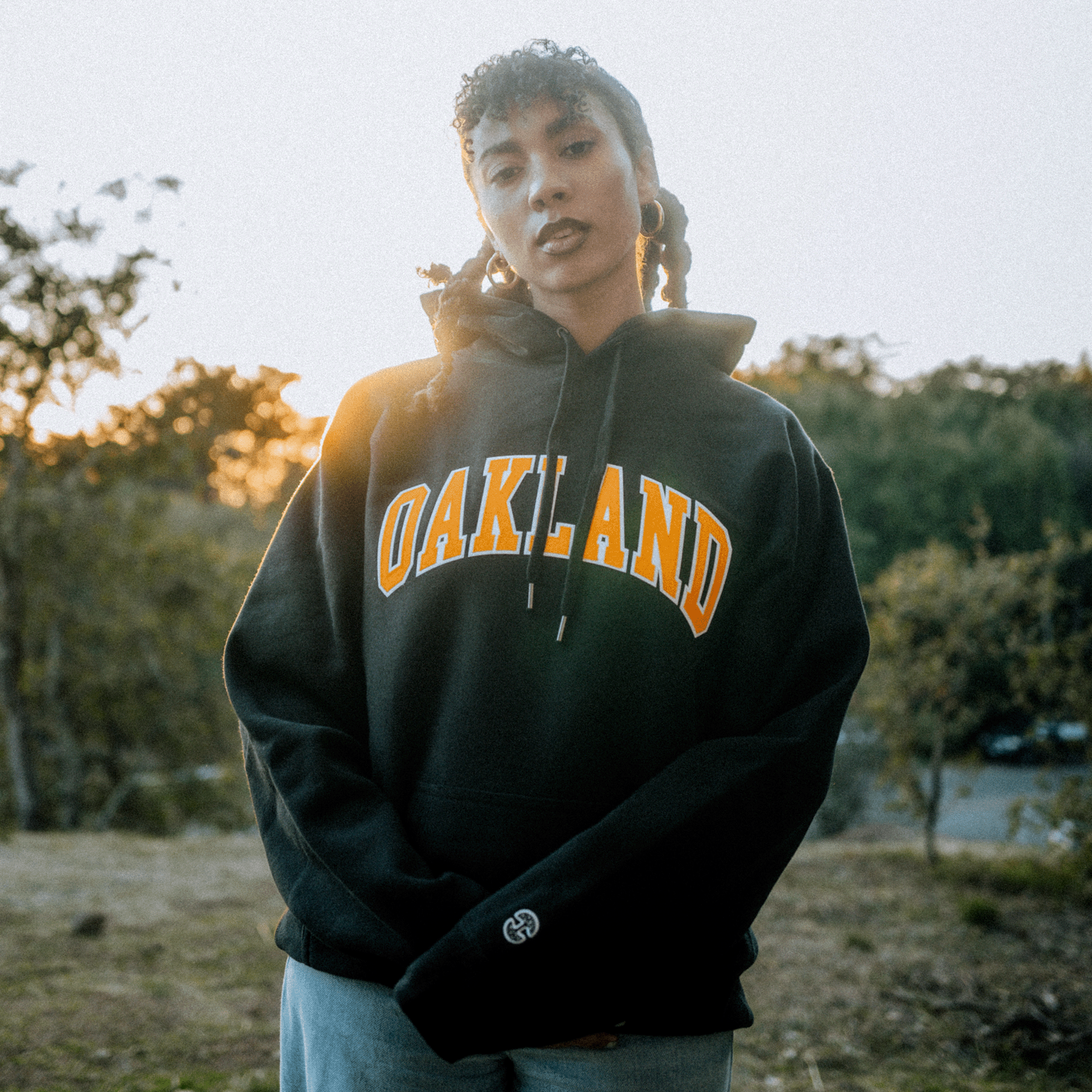 A person with braided hair stands outdoors at sunset, wearing a cozy Alma Mater Heavy Hoodie in black from Oaklandish, featuring "OAKLAND" in bold yellow letters. The fleece provides warmth as the background features trees and a soft, blurred sky, creating a serene ambiance.