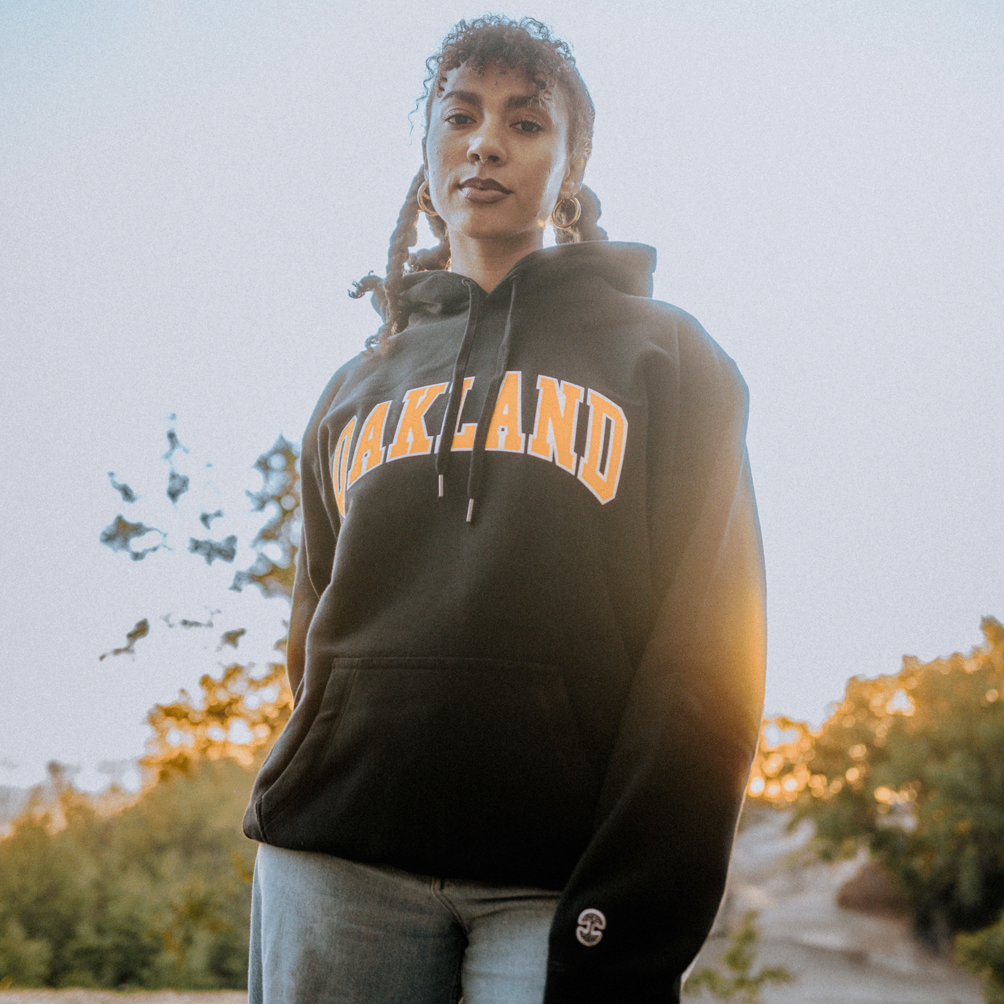 A person with curly hair in braids sports the Alma Mater Heavy Hoodie by Oaklandish, featuring "OAKLAND" in bold yellow letters against a black background. Standing outdoors, they are framed by trees and a bright sky, while the sunlight casts a warm glow around them, emphasizing their confident and relaxed demeanor.