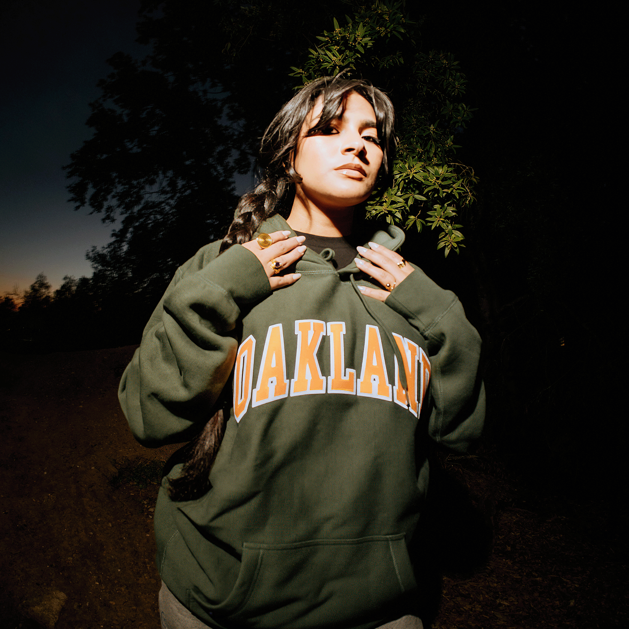 A woman with long braided hair stands confidently in a dimly lit outdoor setting, wearing an Alma Mater Heavy Hoodie from Oaklandish, featuring the word "OAKLAND" prominently in yellow. She accessorizes with multiple rings as trees and a dark sky form the backdrop, creating a dramatic, moody atmosphere.