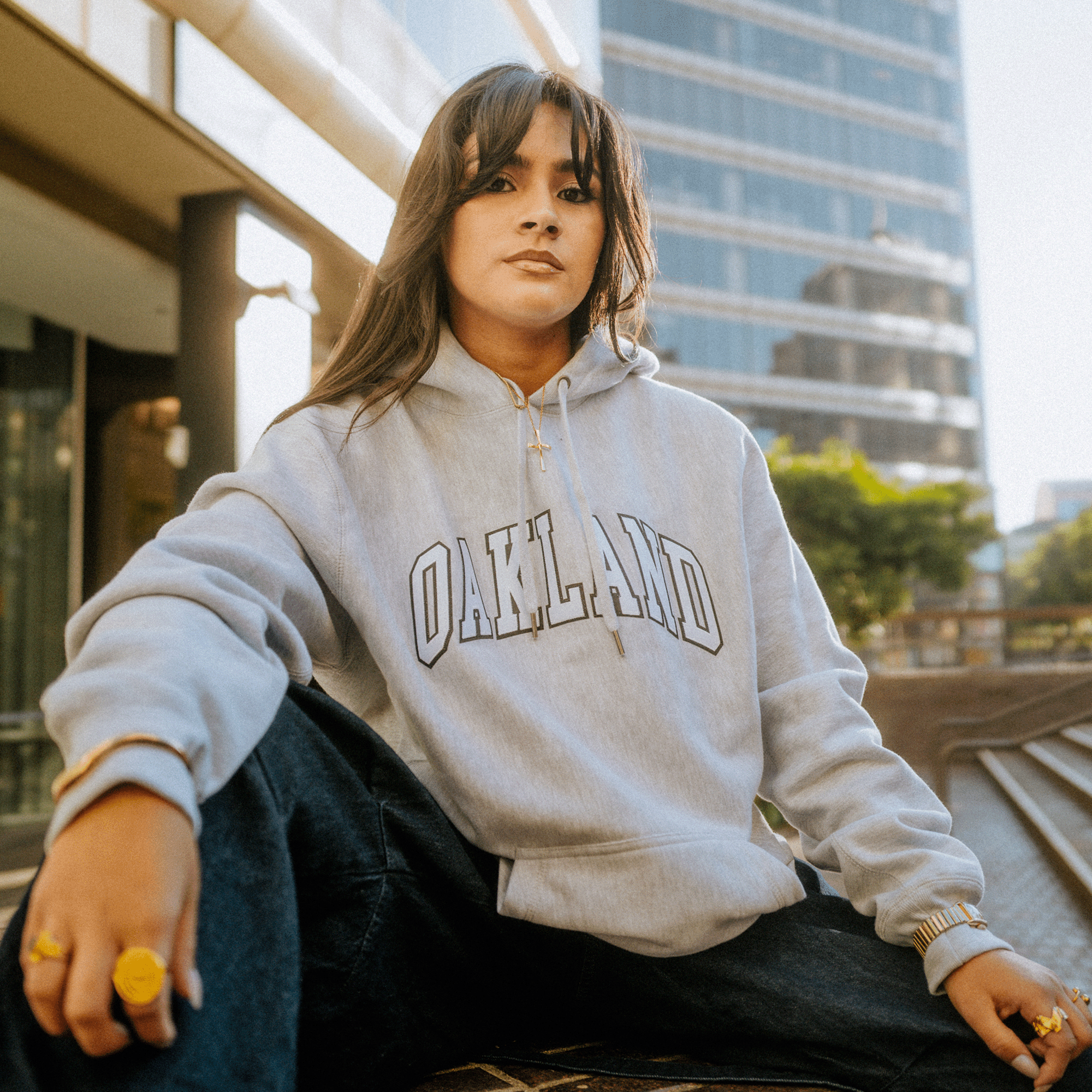 A person wearing the Alma Mater Heavy Hoodie by Oaklandish, featuring "OAKLAND" across it, sits outdoors. Their long hair is framed by modern buildings. With a confident look and one knee raised, hands resting on their legs, the sunlight bathes the scene, suggesting a warm day.