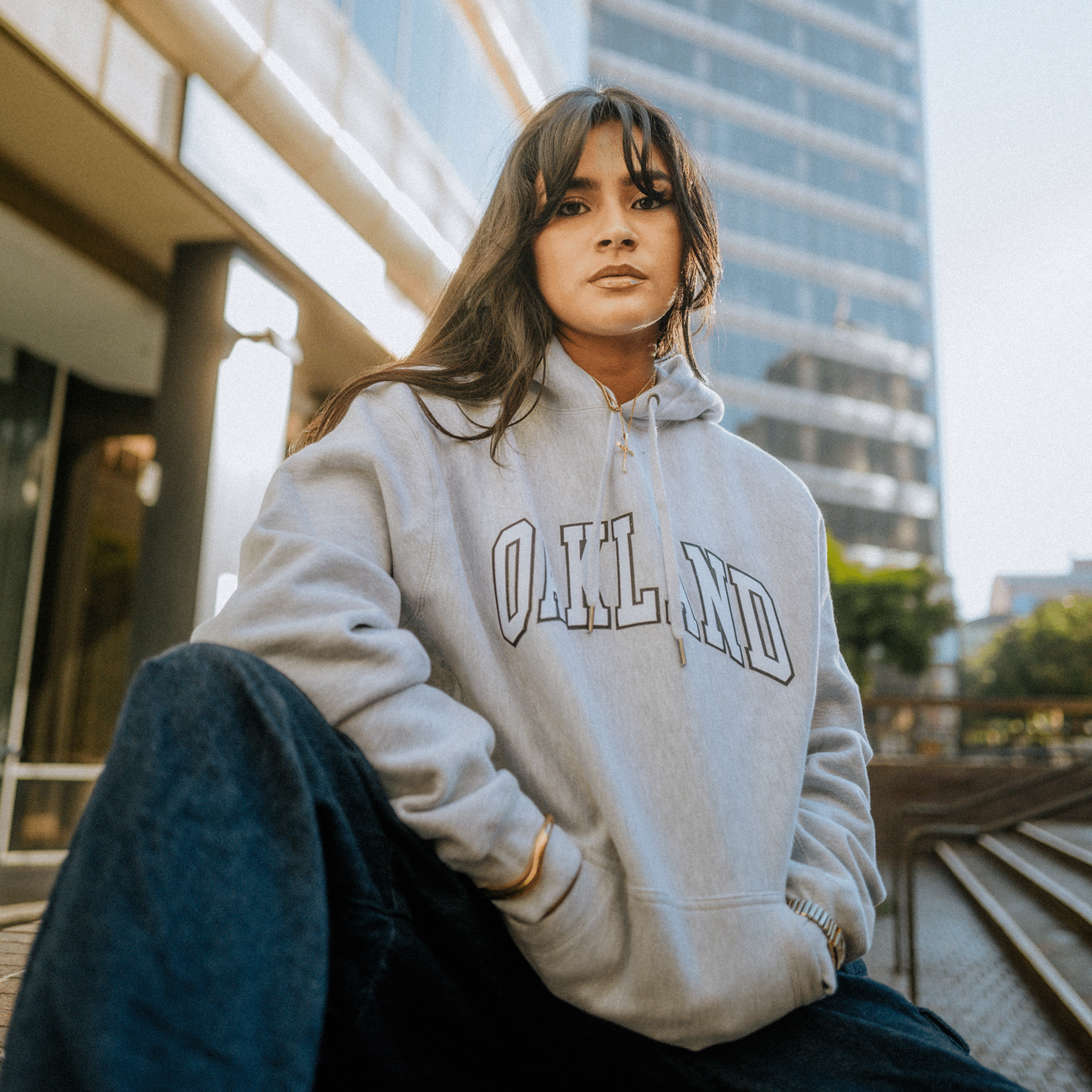 A person with long dark hair sits outdoors, wearing the Oaklandish Alma Mater Heavy Hoodie. With one knee up, they confidently gaze at the camera. In the background, a tall building and trees under a clear sky capture the essence of an urban setting.