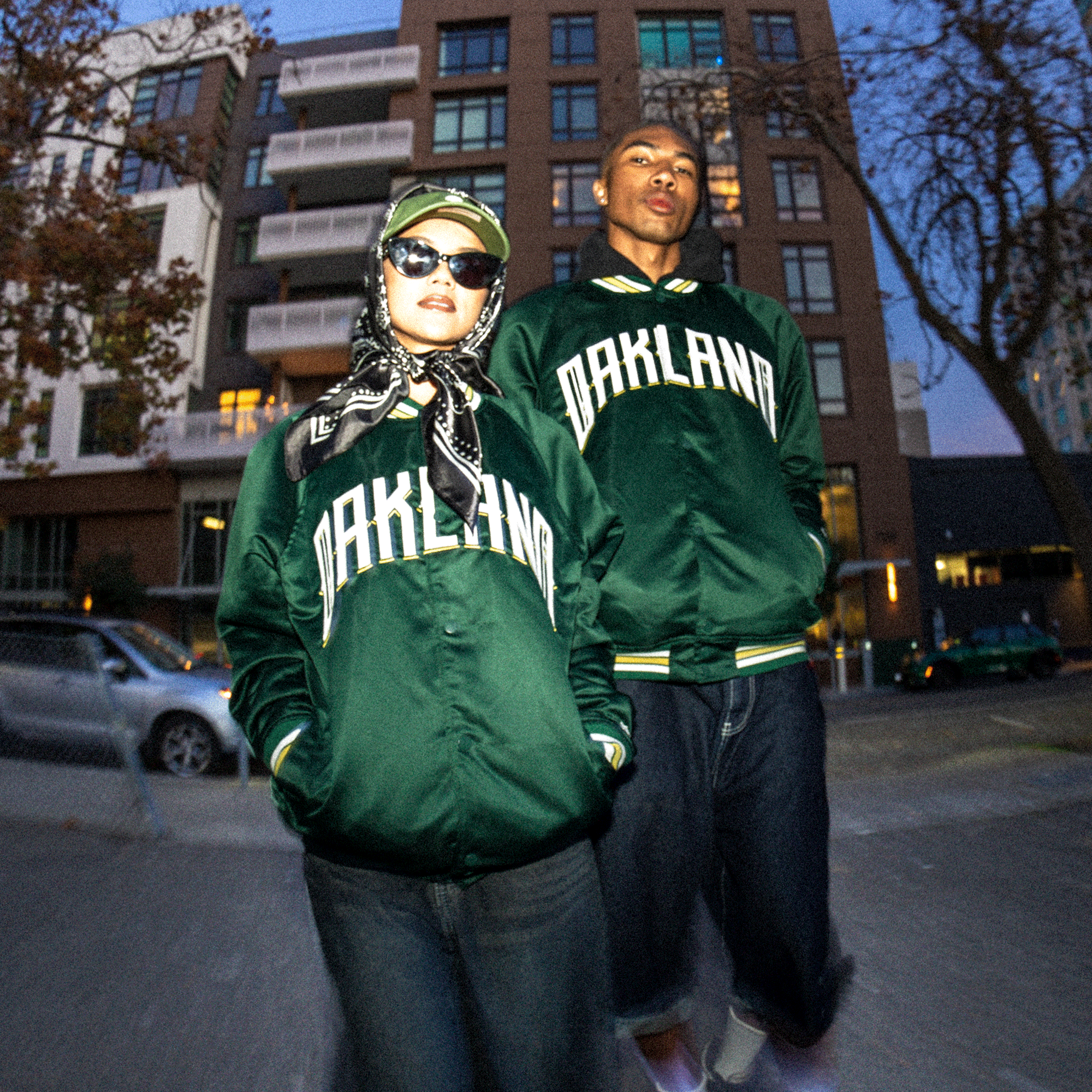 Two people stand outside an apartment building at dusk, each wearing the Oakland Ballers Satin Jacket. They look confidently at the camera, proudly supporting the Oakland Ballers baseball team in matching bomber jackets.