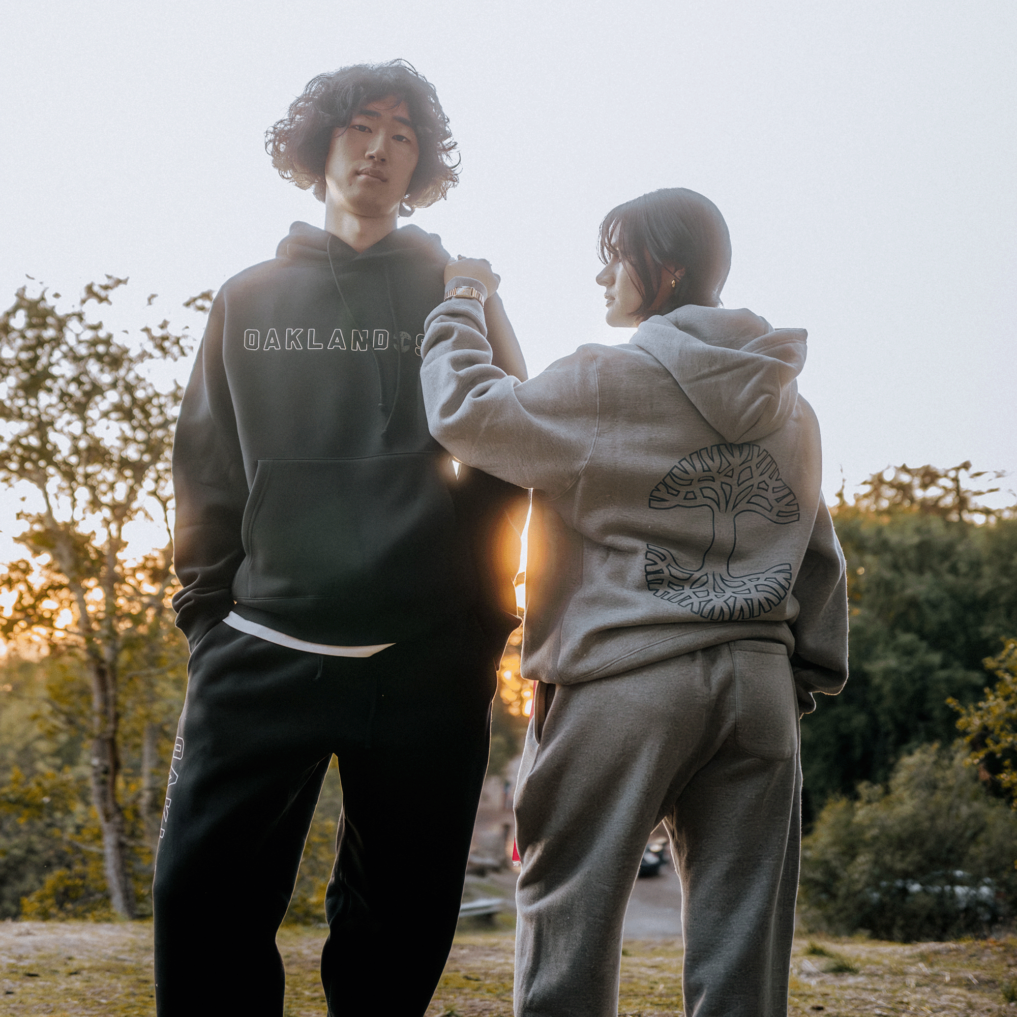 A person in Oaklandish's black "Oakland" hoodie and Bandit Sweatpants stands next to another in a gray hoodie featuring a tree design on the back. They are outdoors, surrounded by trees with a soft sunset in the background. The individual wearing gray gently touches the other's shoulder.