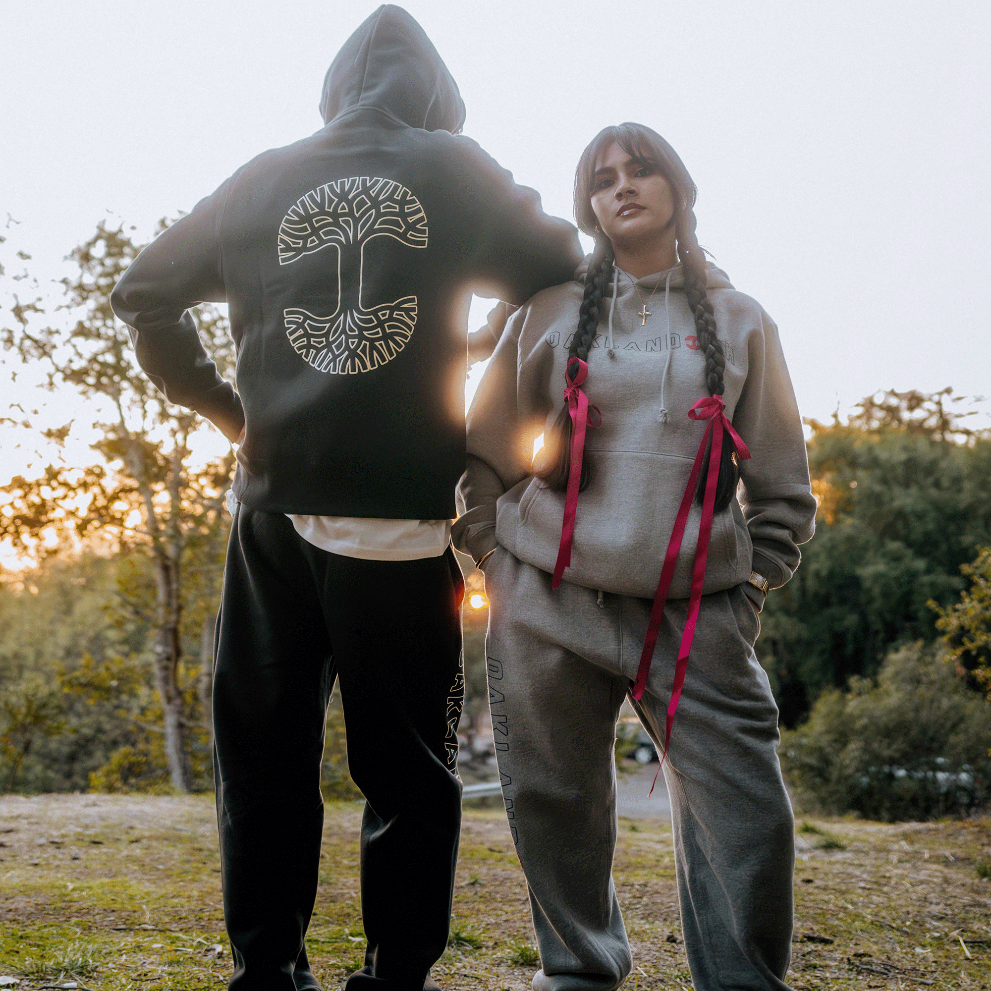 A person wearing the Oaklandish Bandit Hoodie, a black pullover adorned with a large tree design, stands next to someone in a slightly oversized, heather grey hoodie embellished with red ribboned braids. Both individuals are dressed in sweatpants and enjoying the outdoors on a grassy area at sunset, encircled by trees and soft natural light.