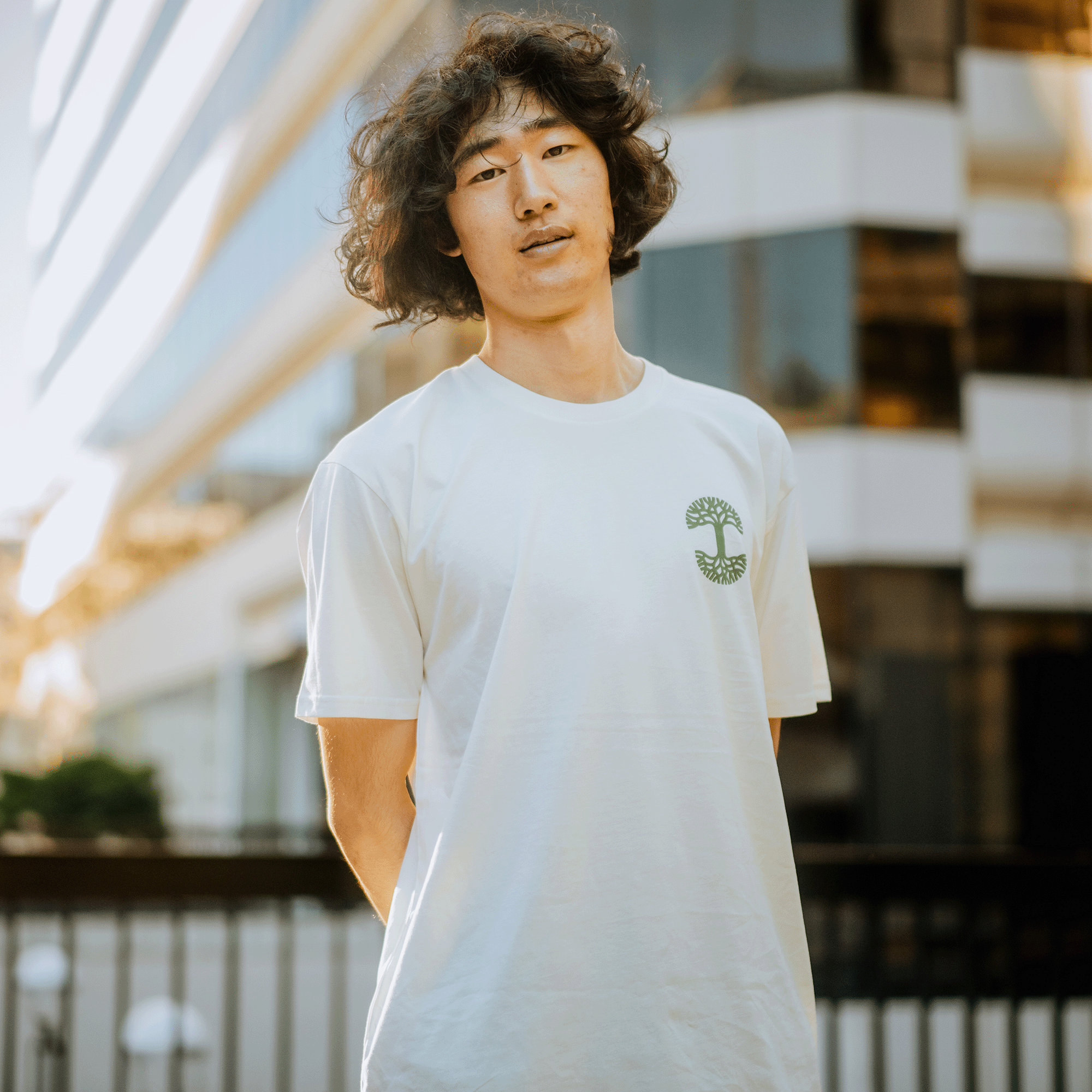 A person with curly hair wearing a white Black Bird Tee from Oaklandish's men's collection, featuring a green logo, stands outside. They have a relaxed expression in front of a modern building with glass and a white facade. The sun shines gently, casting soft warmth over the scene.