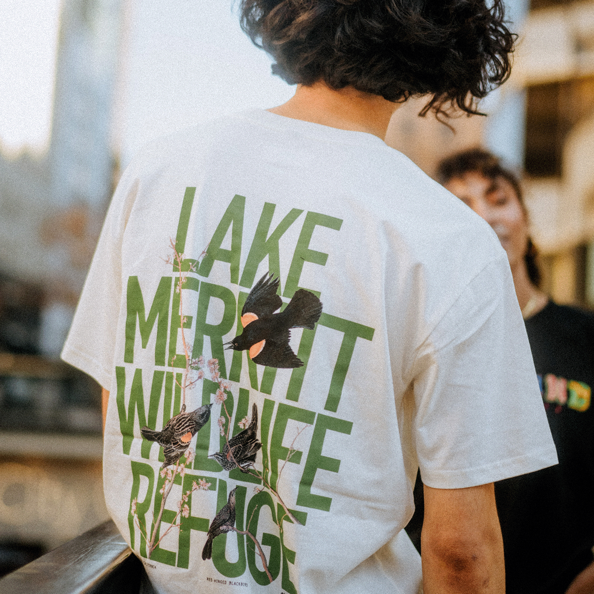 A person with curly hair is wearing the Black Bird Tee from Oaklandish's men's collection, made of 100% cotton. The shirt showcases "Lake Merritt Wildlife Refuge" printed in large green letters on the back, along with illustrations of birds and branches. Another person is blurred in urban surroundings.