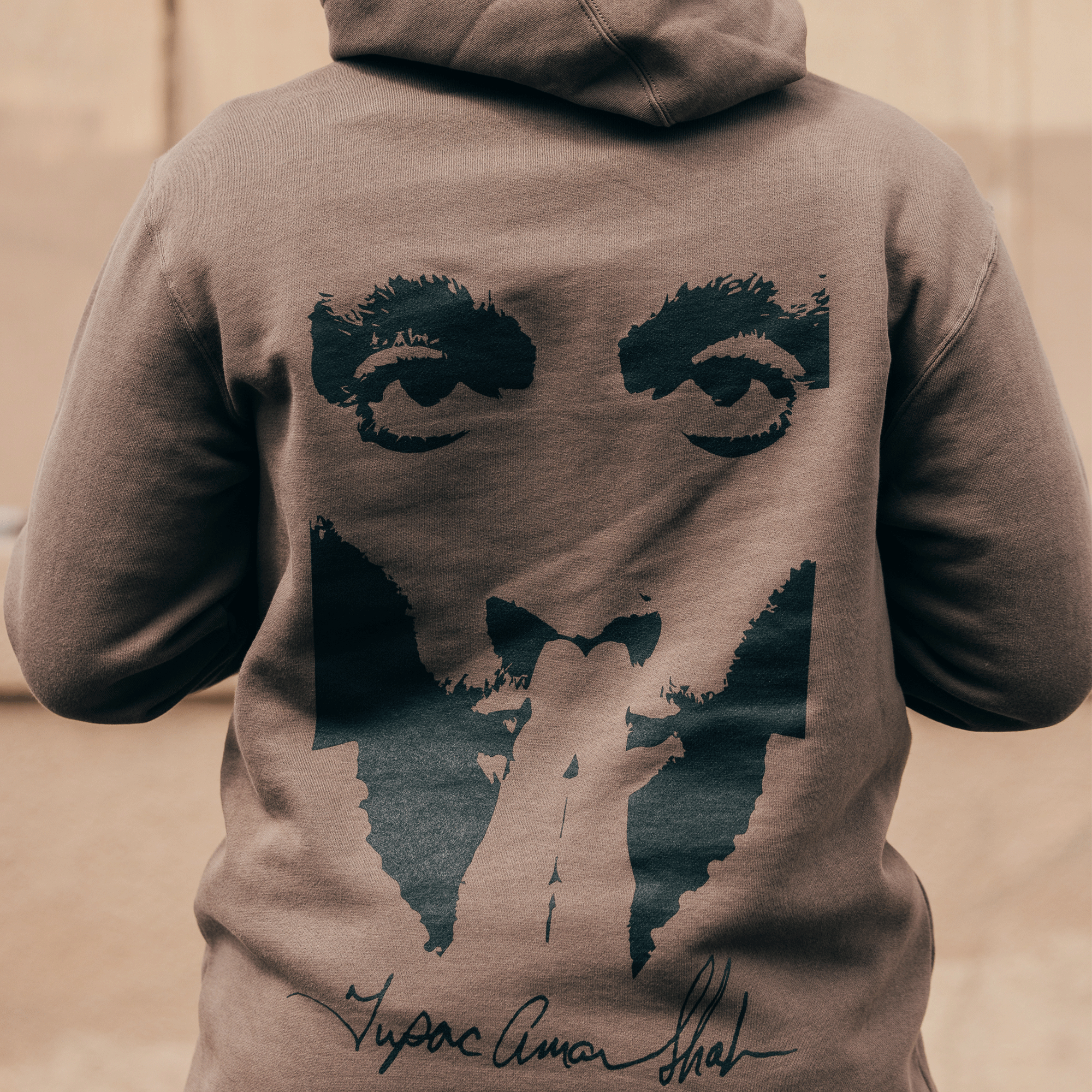 A person is wearing the Bless Hoodie from the Oaklandish x Tupac collection, showcasing a large graphic design on the back with a stylized face of a man with penetrating eyes, facial hair, and the signature "Tupac Amaru Shakur." The background is softly blurred, highlighting the hoodie.