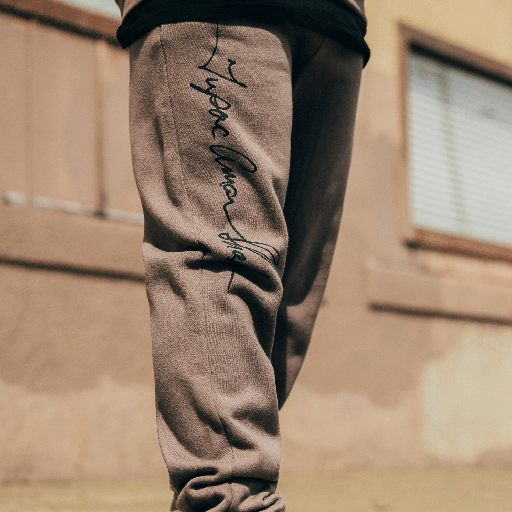 A person stands outdoors wearing the brown Bless Jogger with black cursive writing on the side, part of the exclusive Tupac collaboration by Oaklandish. In the background, there's a building with a window and beige wall. The photo is cropped just above the waist and below the knees, emphasizing this piece from their hip-hop collection.