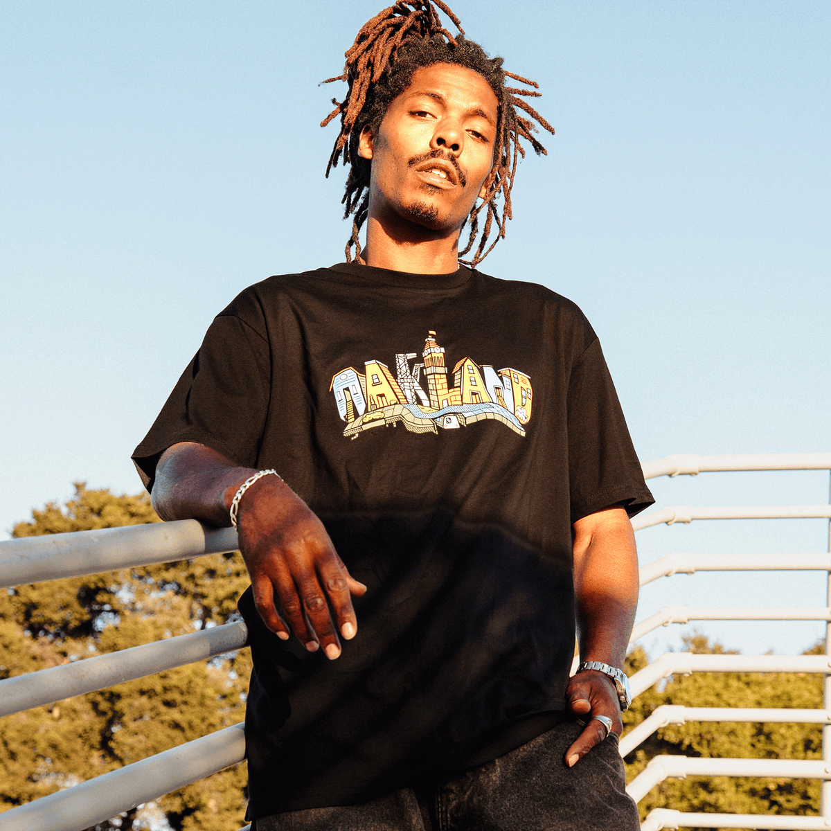 A man with dreadlocks wears a black Oaklandish Blockletters Tee featuring a colorful Oakland cityscape illustration above a stylized bridge, leaning against a white metal railing during golden hour with his hand resting near a silver chain bracelet.