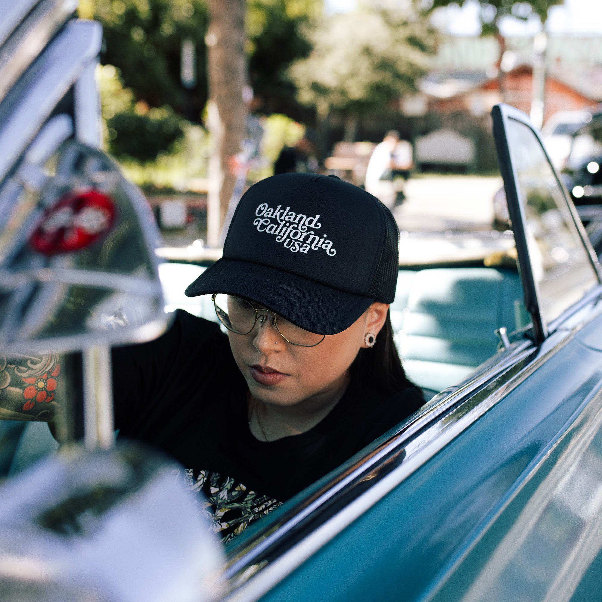 Model wearing a black trucker cap tilted forwardwith foam front panel, mesh back, adjustable back, and white Oakland, California, USA wordmark on the crown while sitting in car.