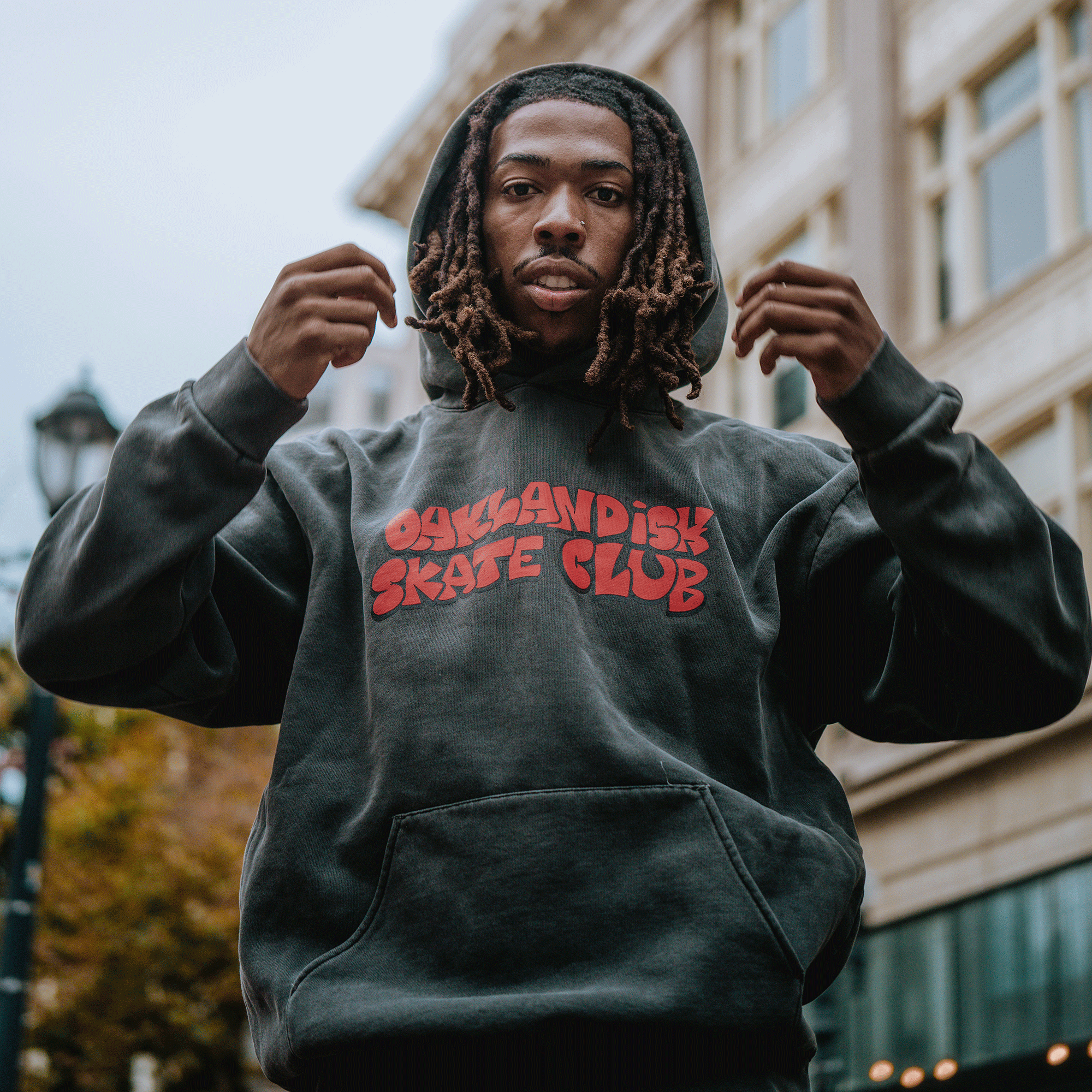A young man with dreadlocks wears the Oaklandish Skate Club Hoodie by Oaklandish, standing outdoors in the city. Embracing skateboard culture, he looks at the camera while holding the hood, with blurred buildings and a streetlamp behind him.
