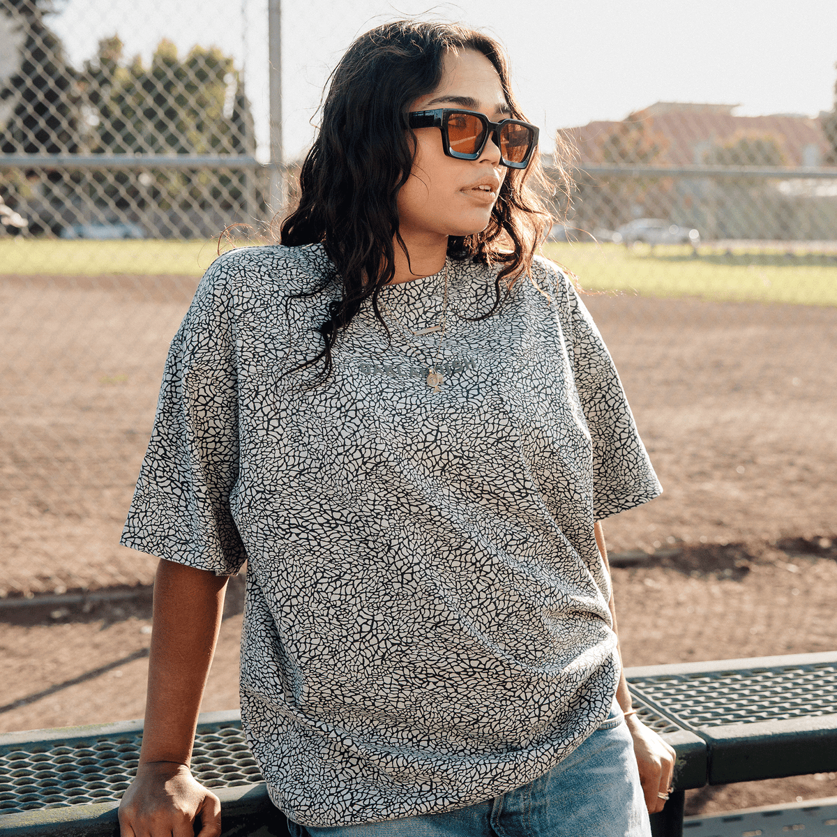 A person with dark, wavy hair and sunglasses wears an Oaklandish Classic Logo Tee in grey, crackle-patterned cotton and light-wash jeans, leaning against a green metal bench on a dirt baseball field as warm sunlight glows behind them.