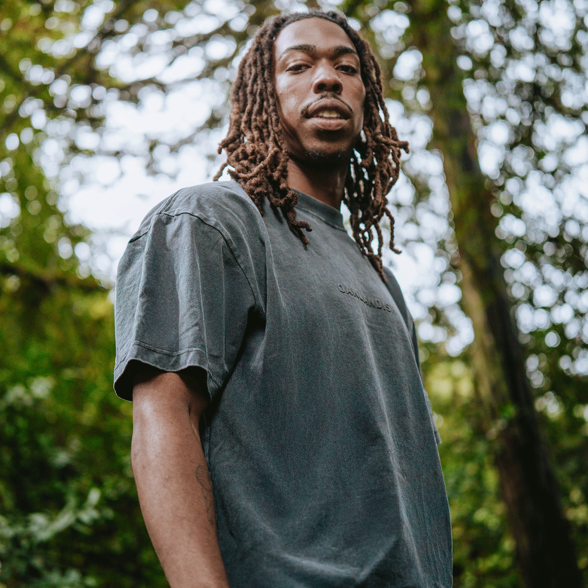 A young man with long, brown dreadlocks stands outdoors amid blurred green foliage, wearing the Oaklandish Classic Logo Tee by Oaklandish. He looks slightly down at the camera in a forest or park-like setting.