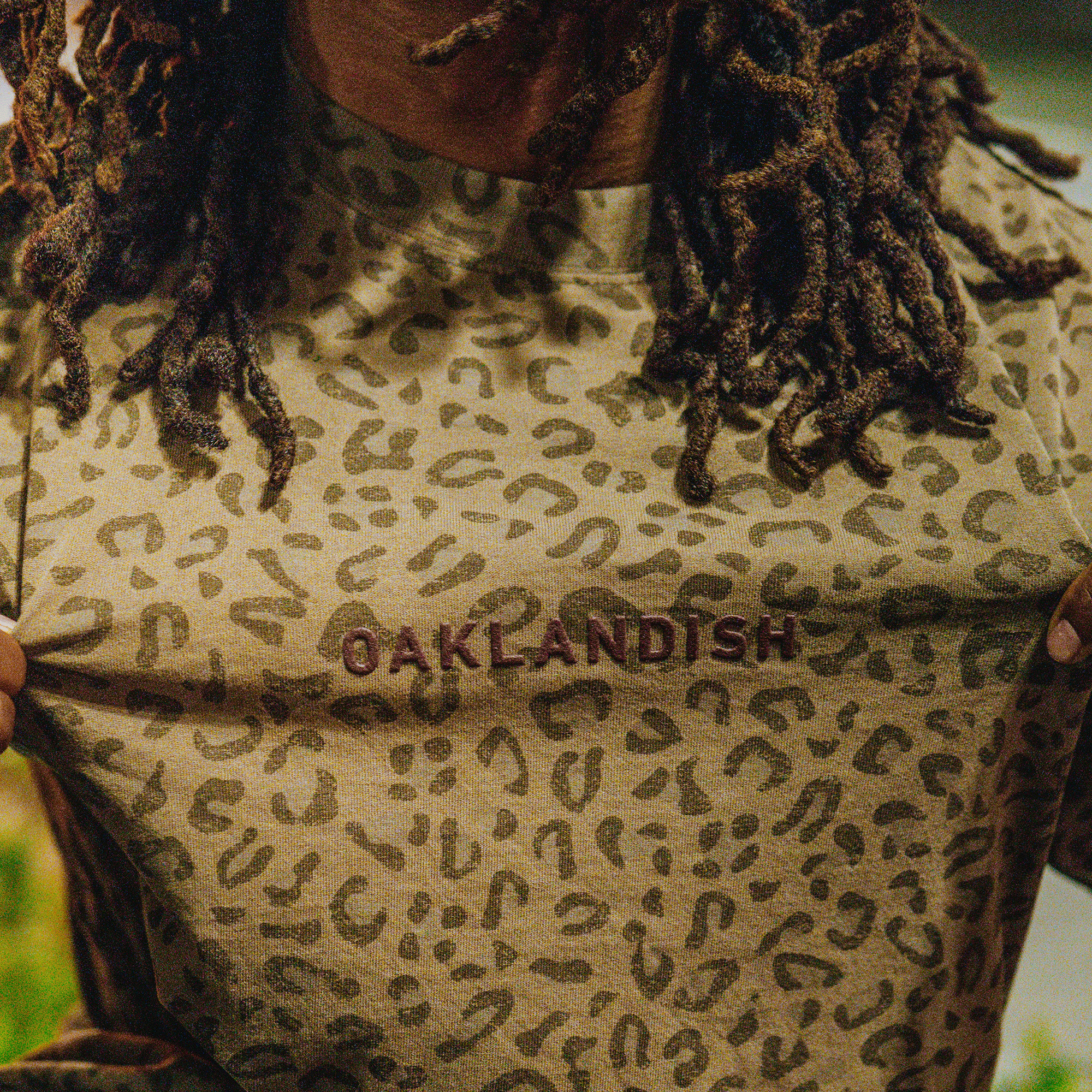 A person with long, curly hair holds out a tan Oaklandish Classic Logo Tee, a men's cotton t-shirt with brown leopard print and "OAKLANDISH" embroidered in dark red across the chest; background is blurred for focus.
