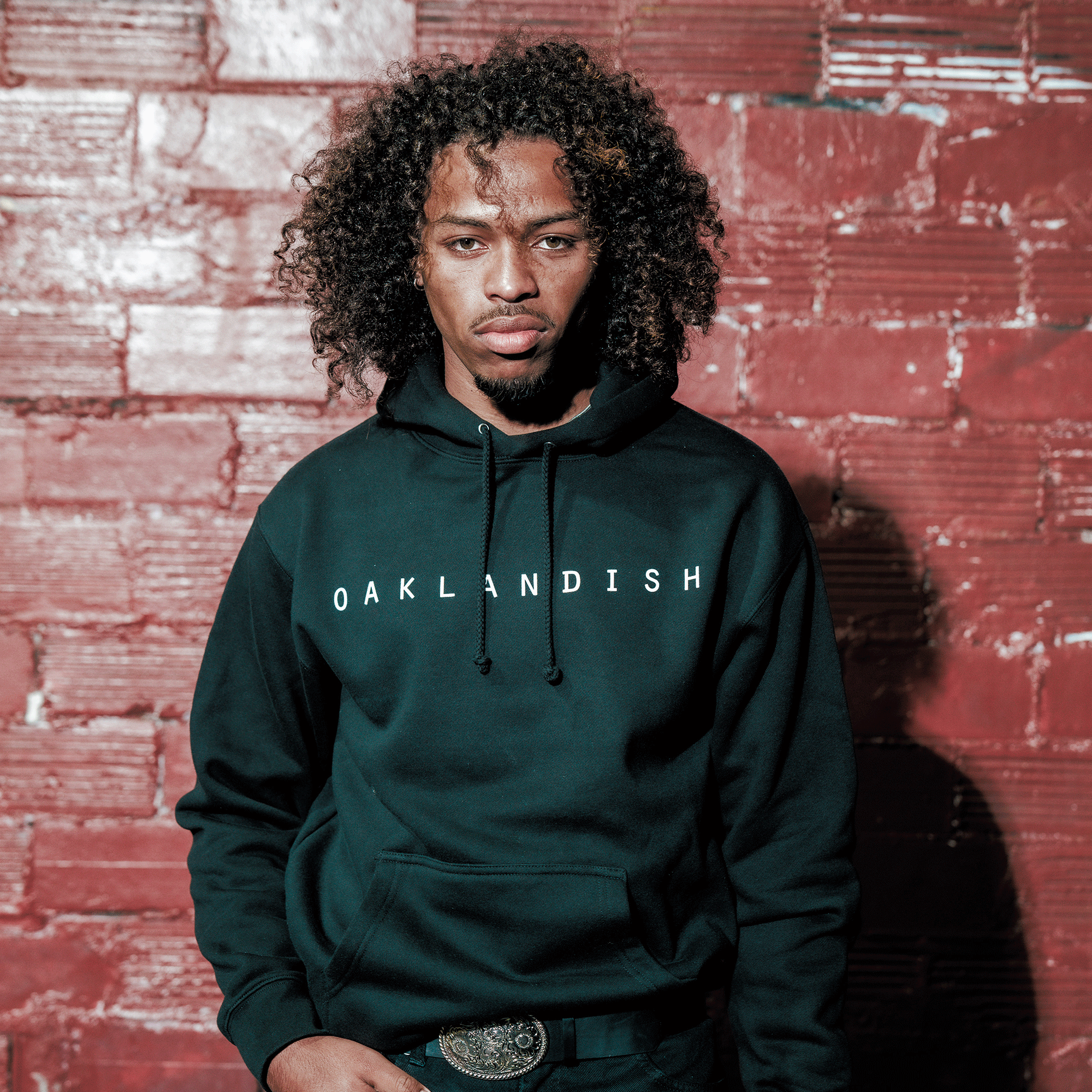 A man with curly hair wears an Oaklandish Classic Hoodie by Oaklandish, standing against a red brick wall with a serious look. Sunlight highlights his face and hair, and his belt buckle is visible below the hoodie.