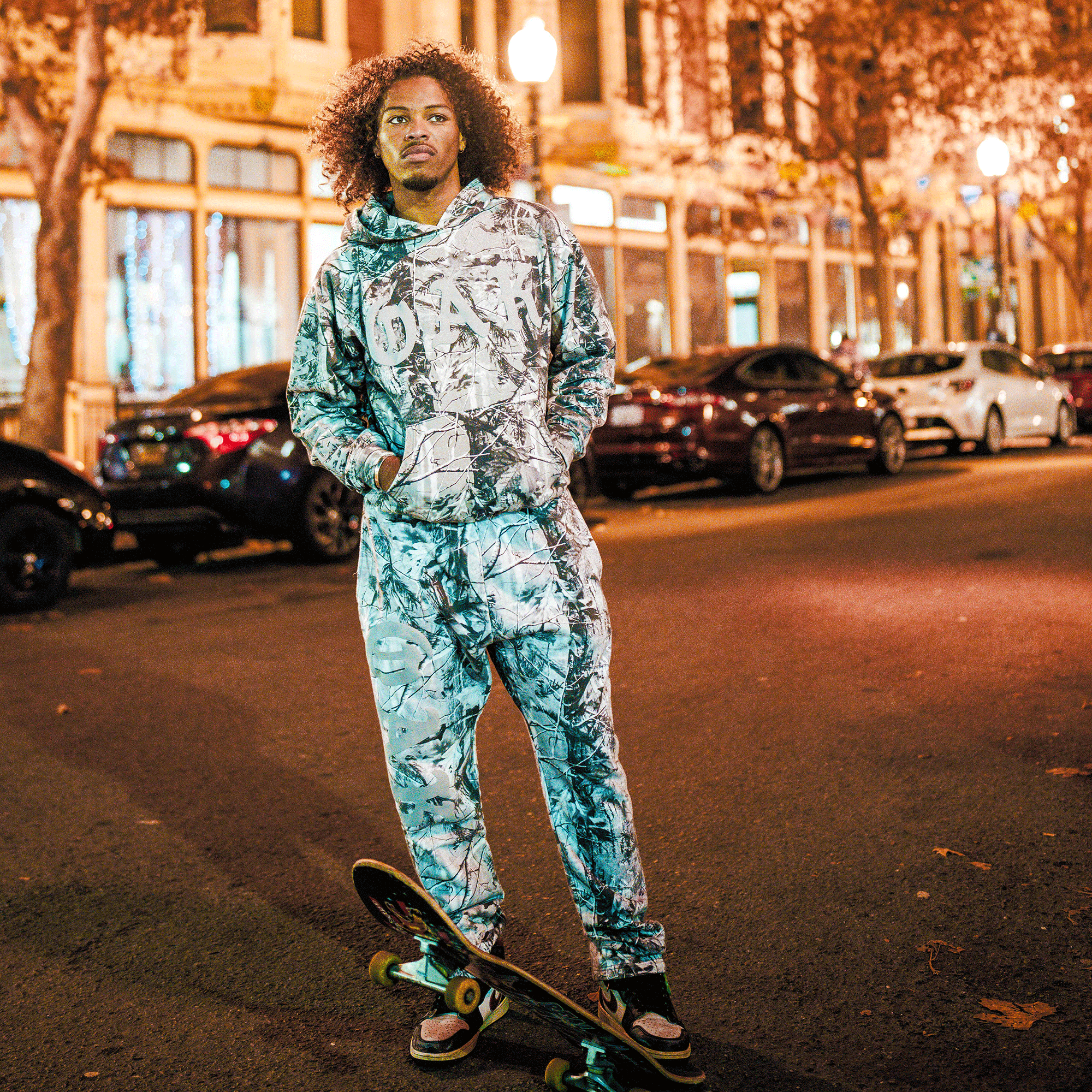A young man with curly hair stands on a city street at night, hands in pockets, wearing the Oaklandish Cork Hoodie and sweatpants, with a skateboard nearby and city lights illuminating parked cars and buildings behind him.