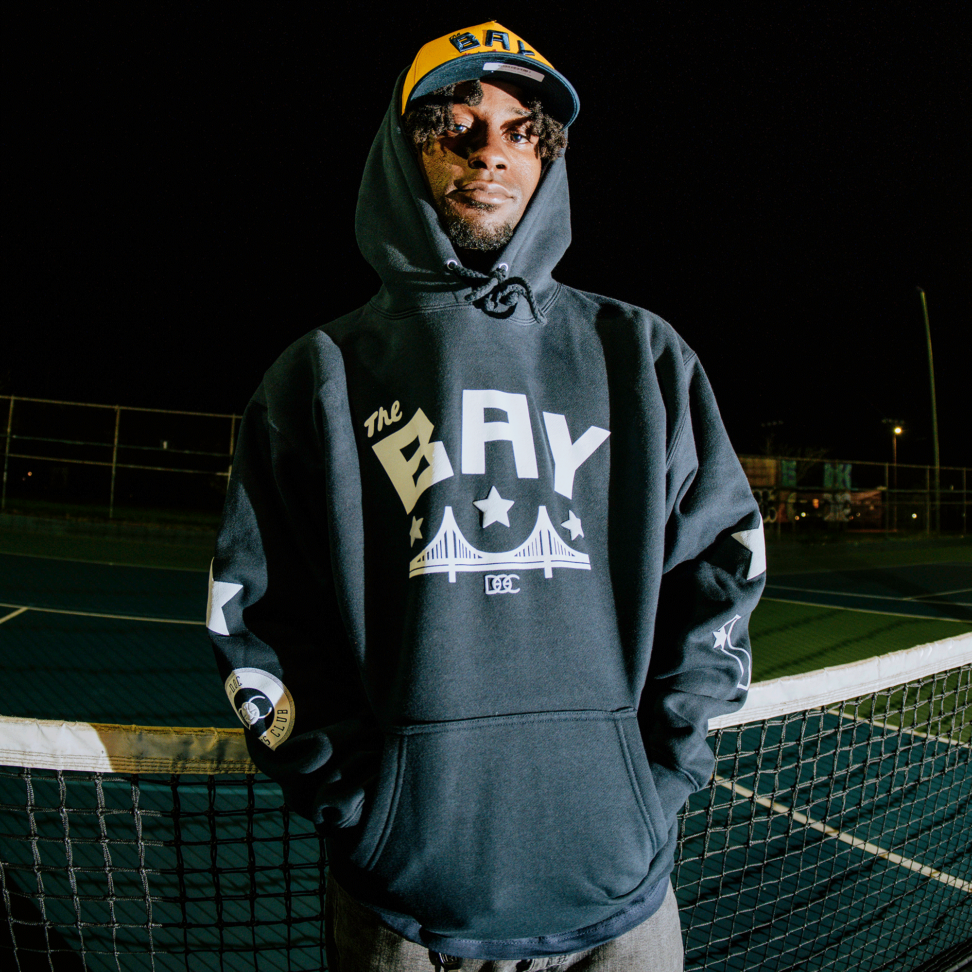 A man in sunglasses and an Oaklandish Bay Bridge Hoodie by DOC, featuring “The BAY” and bridge graphics, with a matching yellow-blue cap, stands on an outdoor tennis court at night under bright lights.