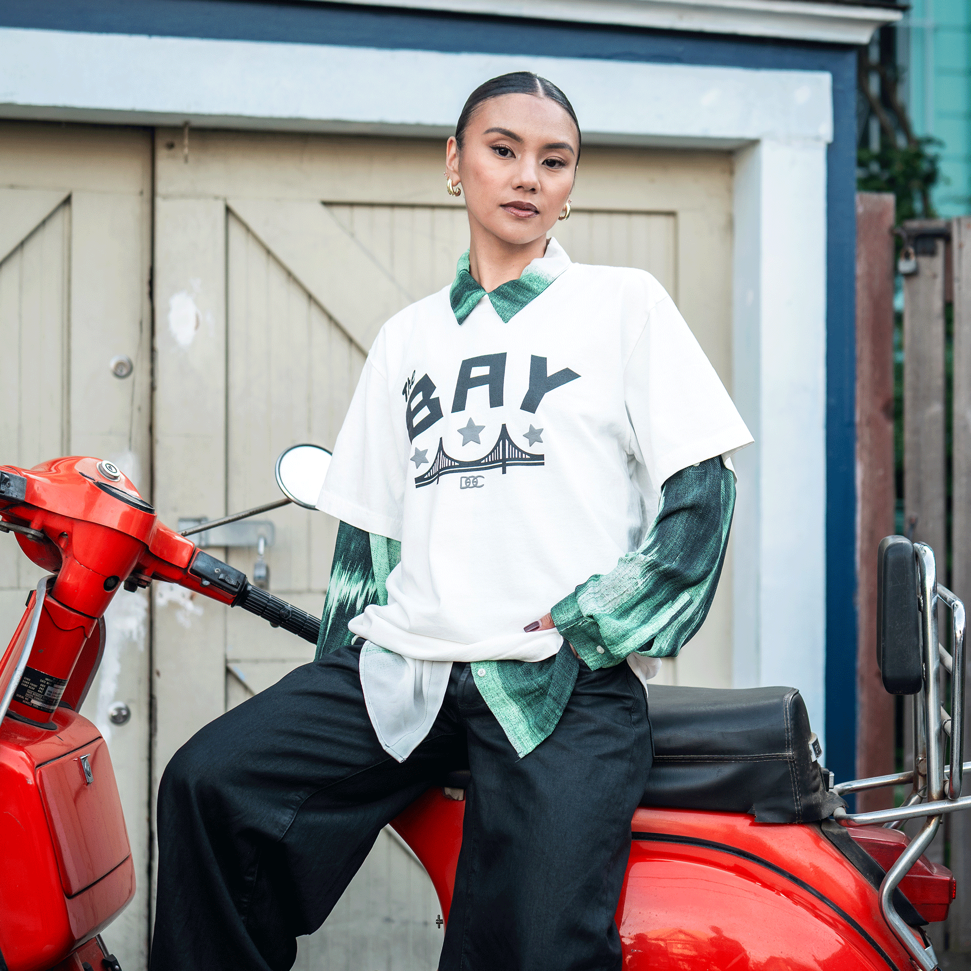 A person with slicked-back hair and hoop earrings poses confidently on a red scooter, wearing the Bay Bridge Tee By DOC V2 from Oaklandish over a green collared shirt and loose black pants, in front of beige double doors with blue trim.