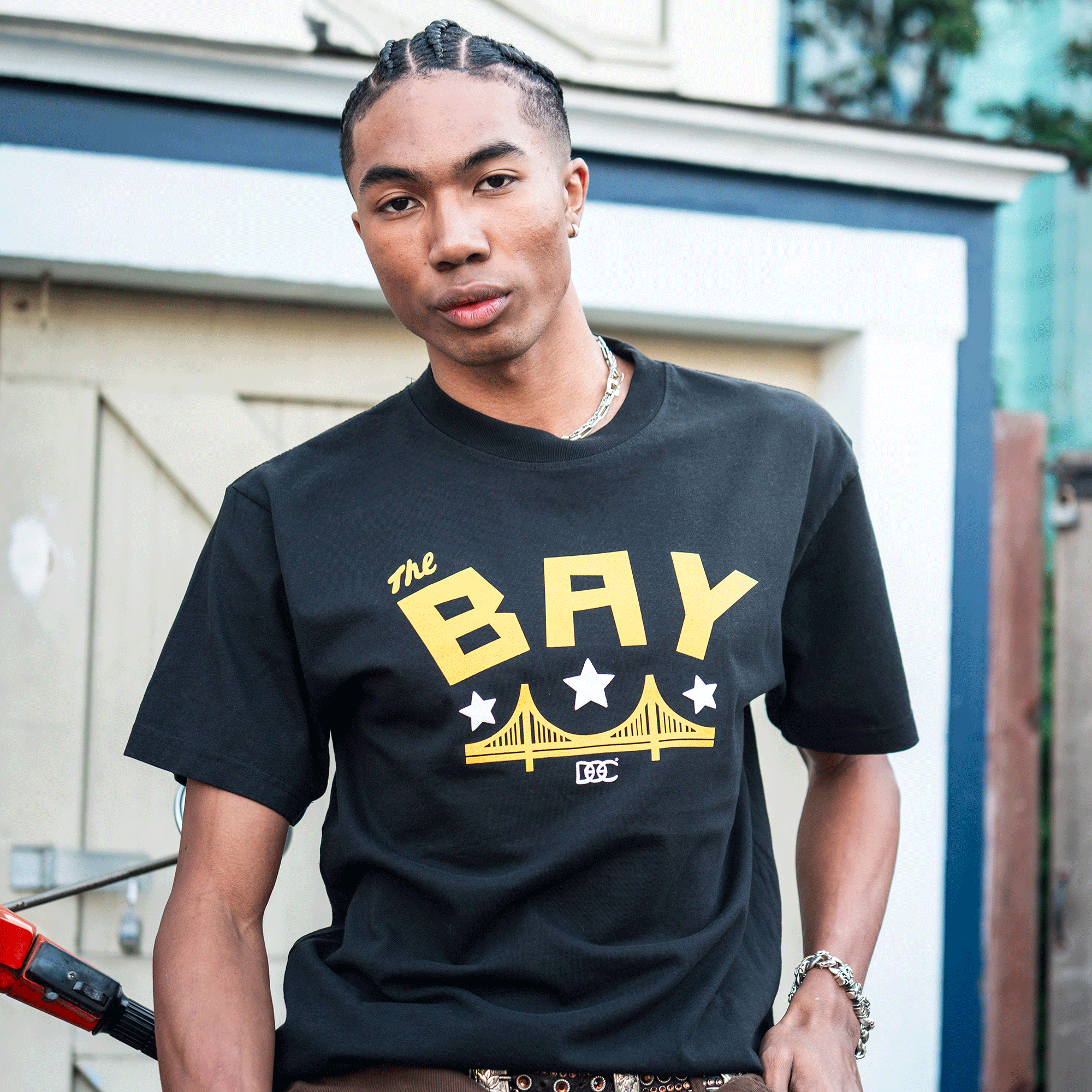 A young man with braided hair stands outside wearing the Bay Bridge Tee By DOC V2 by Oaklandish. The black heavyweight cotton men’s shirt features bold yellow “The BAY” text, a suspension bridge, and stars. A bike handlebar is in the foreground.