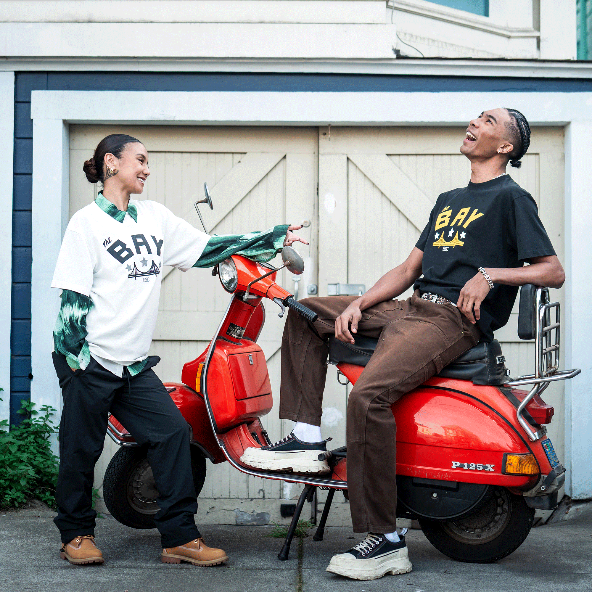Two young adults laugh by a red Vespa. The woman layers her Bay Bridge Tee By DOC V2 from Oaklandish over a green shirt, while the man, in the same tee, sits on the scooter outside a garage.
