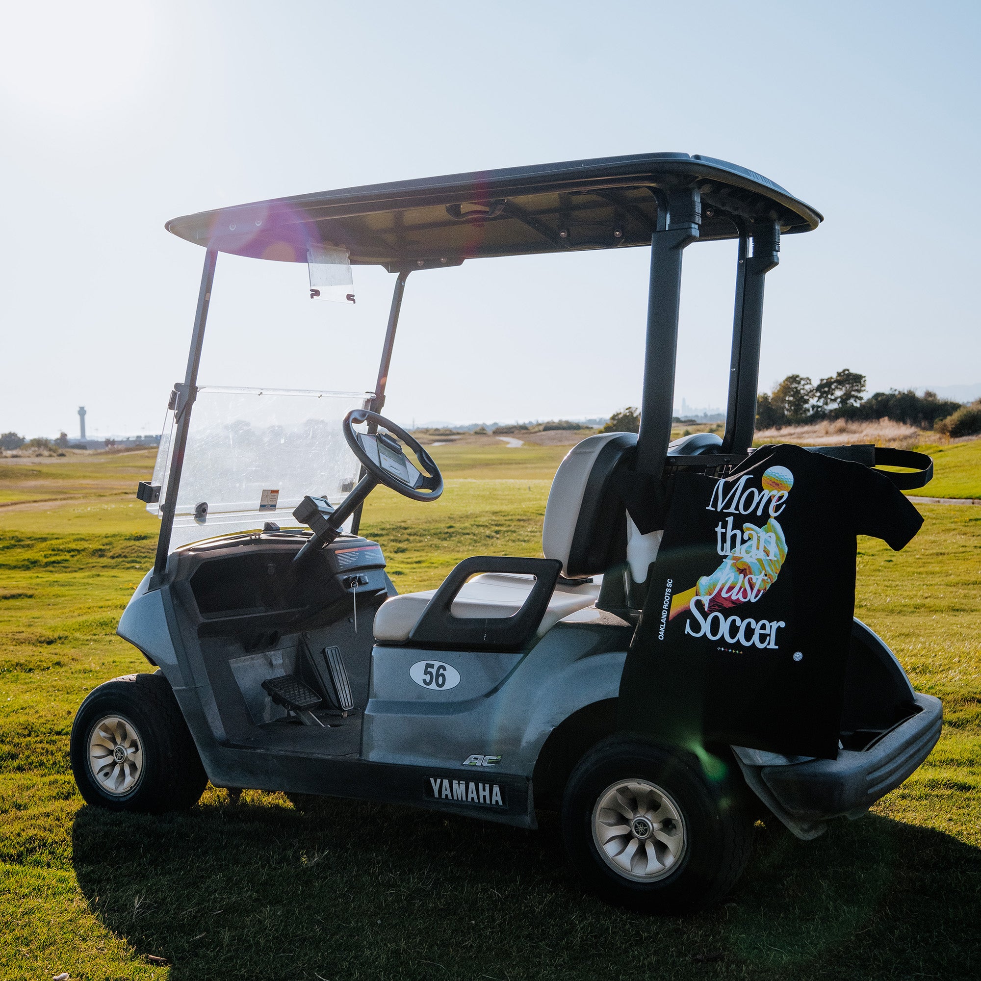 A Yamaha golf cart labeled with the number "56" is parked on a grassy area under a clear sky. An Oakland Roots SC More Than Soccer Tee, showcasing a colorful design and the text "More than just Soccer," is draped over the back of the seat. In the distance, there is a lighthouse-shaped structure.