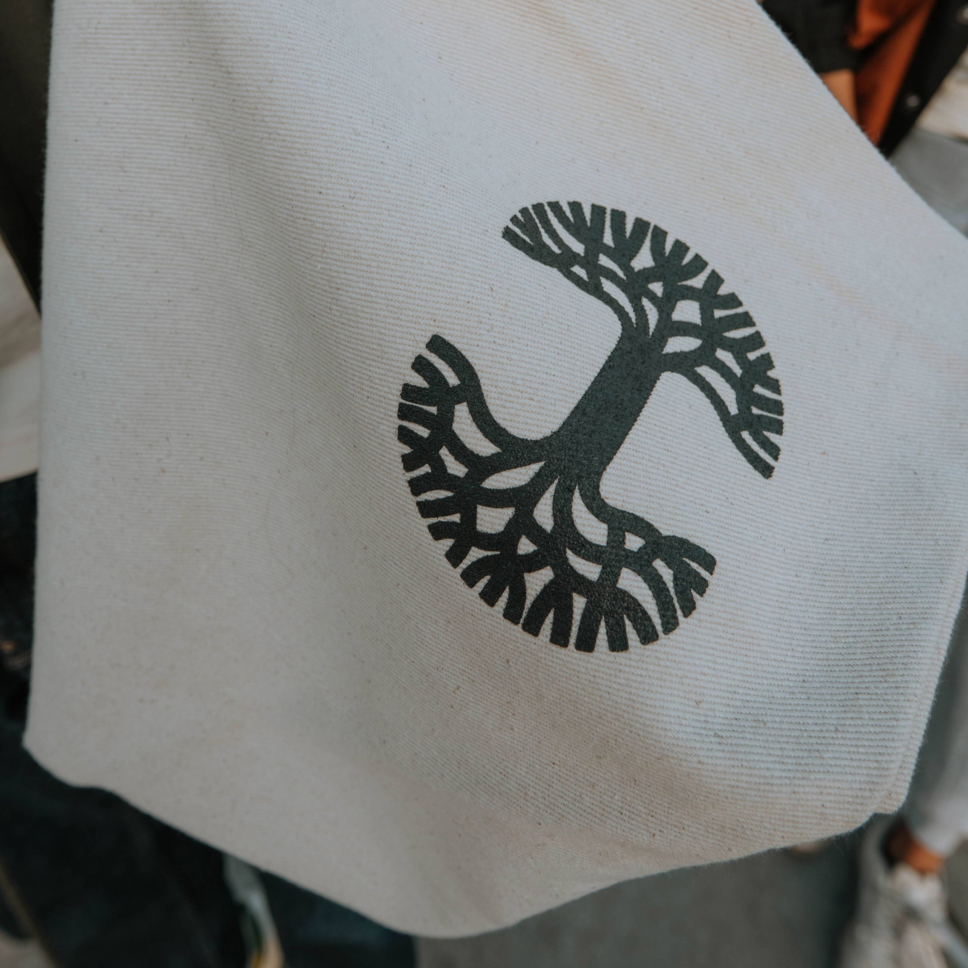 Close-up of an Oaklandish Ultra Canvas Tote showcasing a black stylized tree design. This design includes a central trunk with symmetrical roots and branches. The 100% cotton tote bag is partially shadowed, with a blurry backdrop hinting at an outdoor scene.