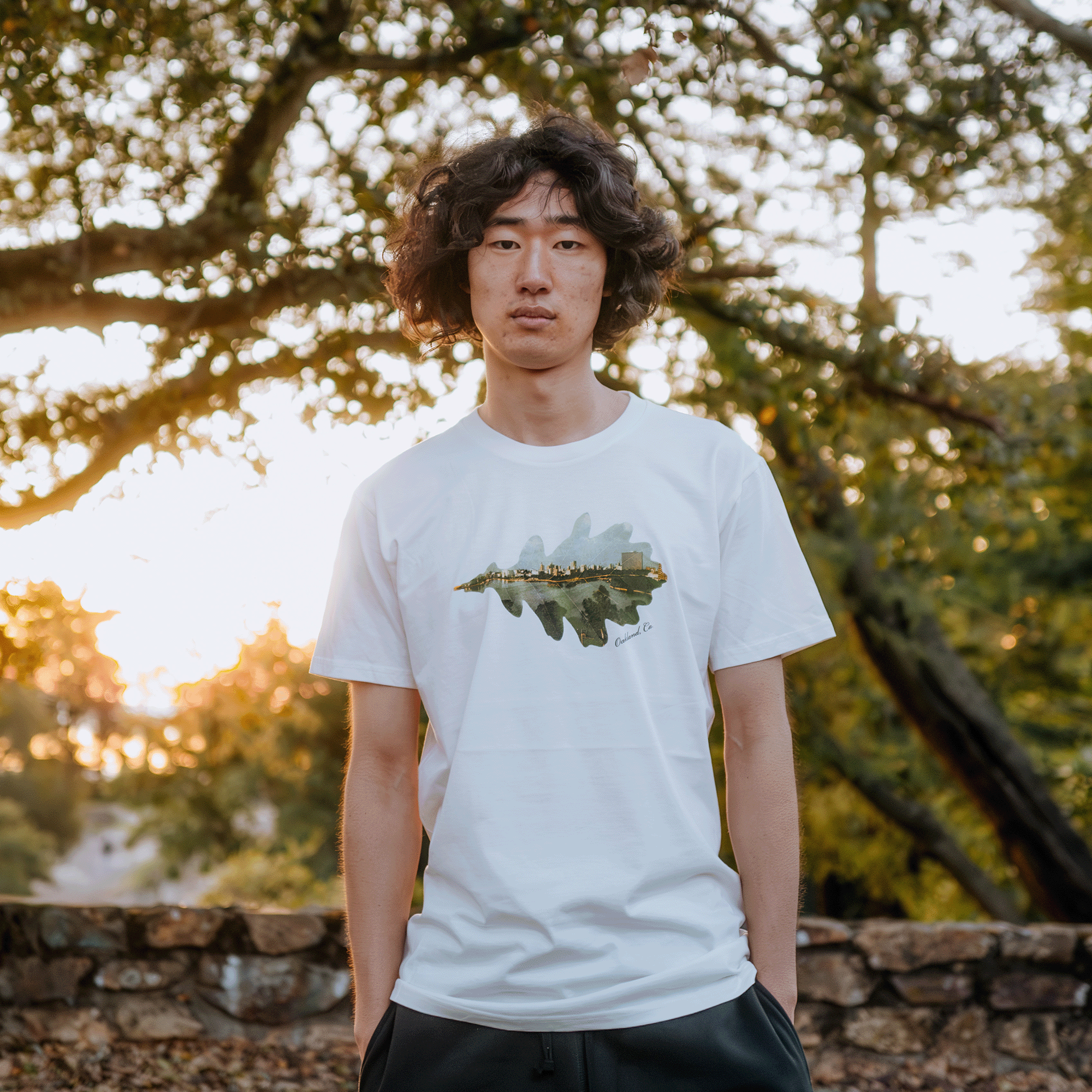 A person with curly hair stands outdoors, wearing a Double Exposure Tee from Oaklandish. This classic fit white T-shirt is made of 100% cotton and features a nature-themed design. They are in a park with trees and sunlight filtering through the leaves, creating a warm ambiance. The background includes a low stone wall and greenery.