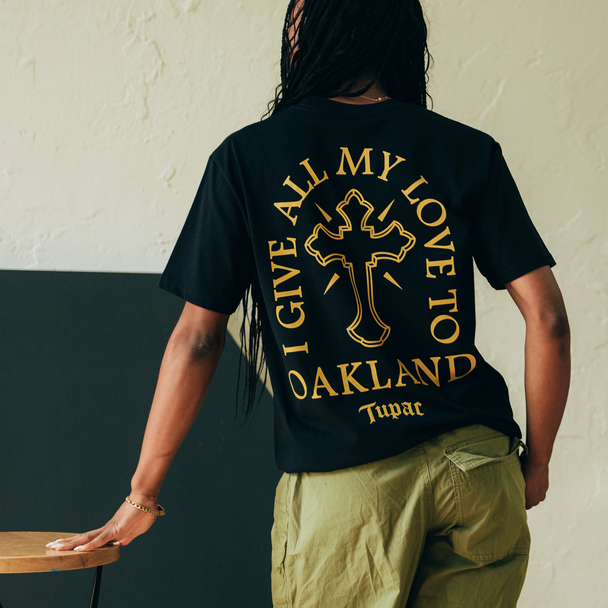 A person with long braided hair stands in front of a beige wall, emanating hip-hop vibes in a black "Double Cross Tee" by Oaklandish, featuring yellow lettering that reads "I give all my love to Oaklandish" and a cross design. They pair it with green pants while standing near a wooden table.