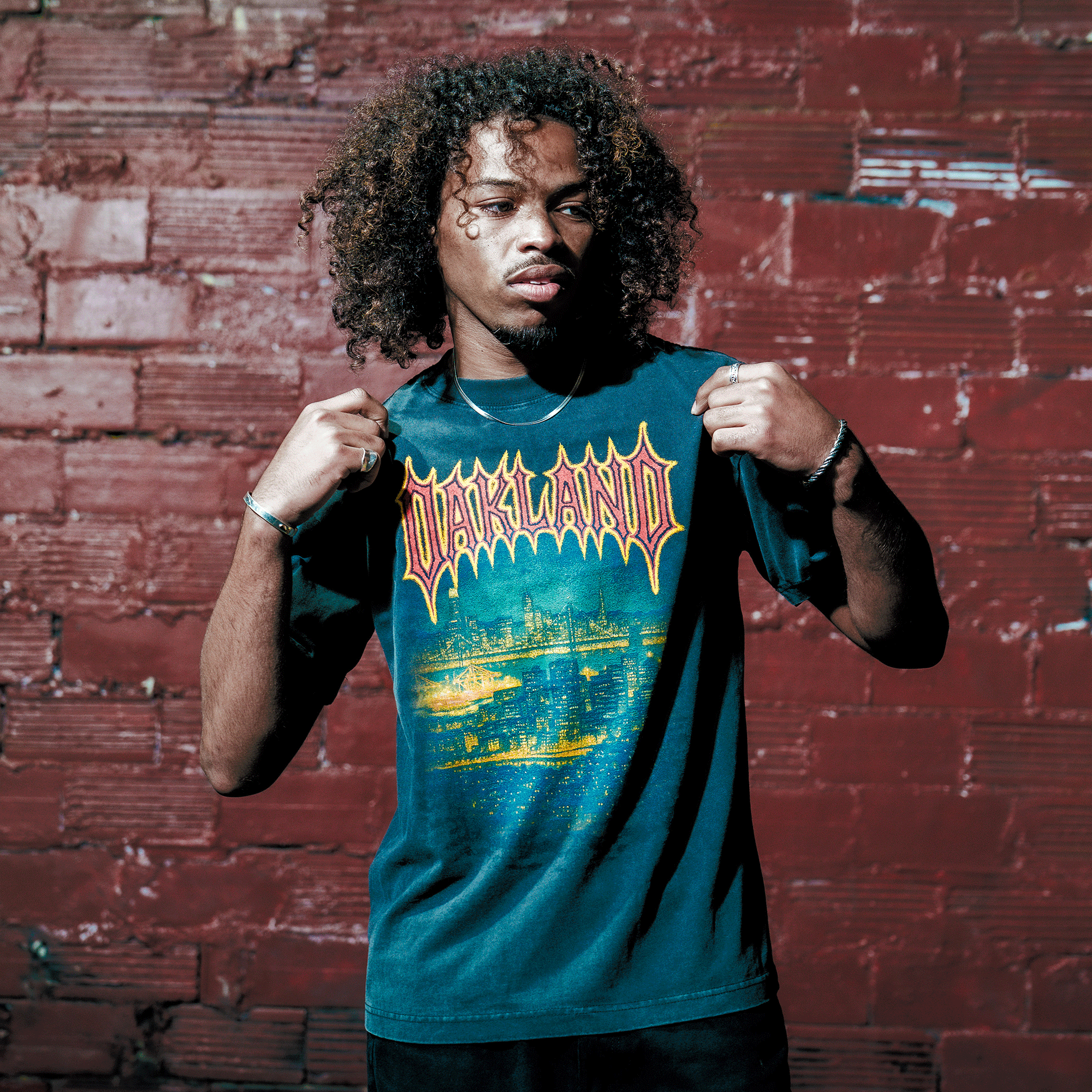 A young man with curly hair stands by a worn red brick wall, holding out the sides of his Oaklandish Ember Tee—a classic fit men's black t-shirt made from 100% cotton, featuring "OAKLAND" and a vibrant cityscape graphic.