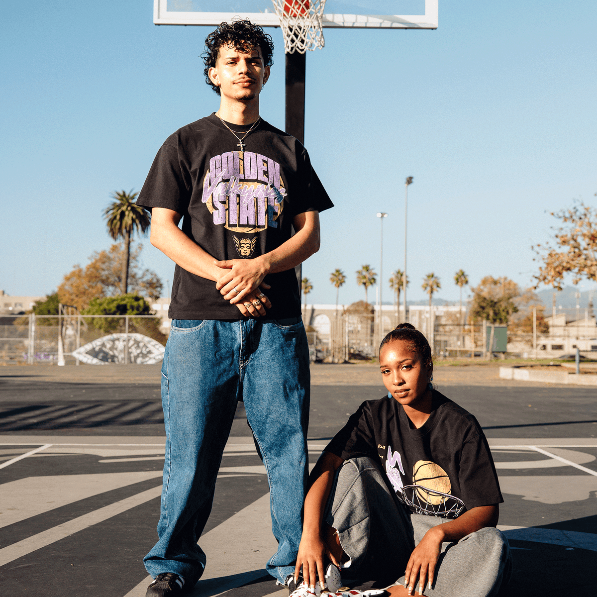 A man and a woman in classic fit GSV Fade Away Tee shirts pose on a paved basketball court; the man wears blue jeans and a cross necklace, the woman wears grey sweatpants, and both are framed by a basketball hoop against a clear blue sky.