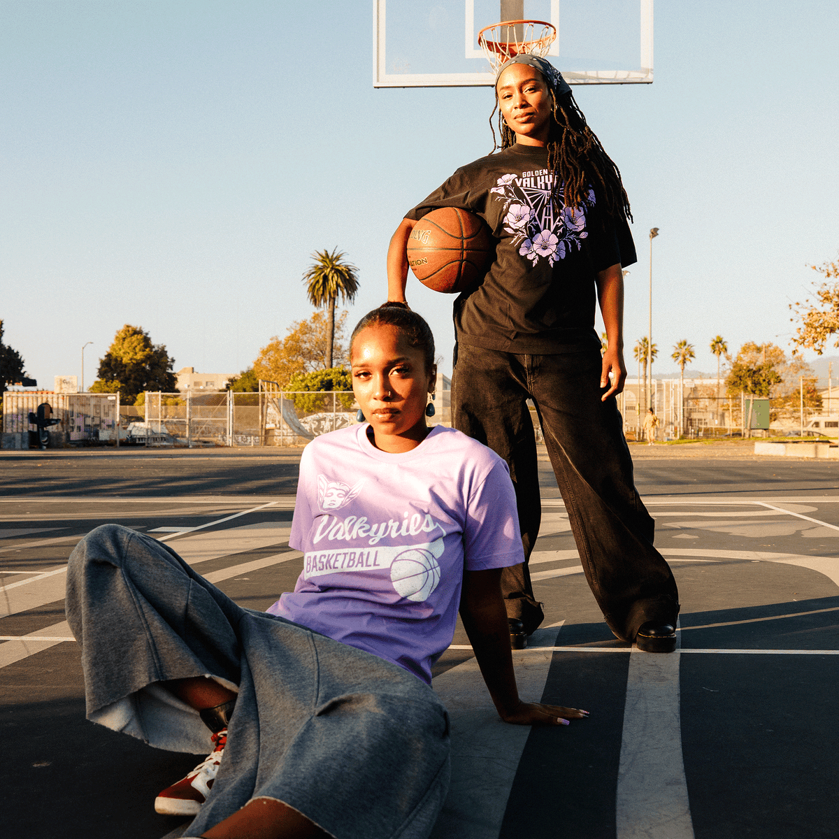 Two Black women pose on an outdoor basketball court during golden hour; one sits on the ground wearing a GSV Poppies Tee and wide-leg jeans, while the other stands behind her in a black graphic t-shirt, holding a basketball.